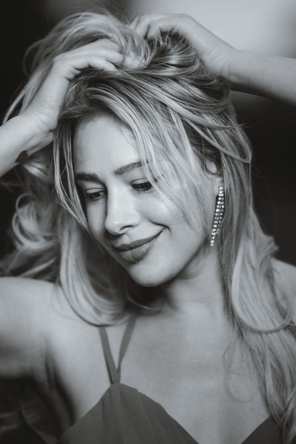 Black and white photo of a blonde woman styling her hair up while wearing dangling earrings.
