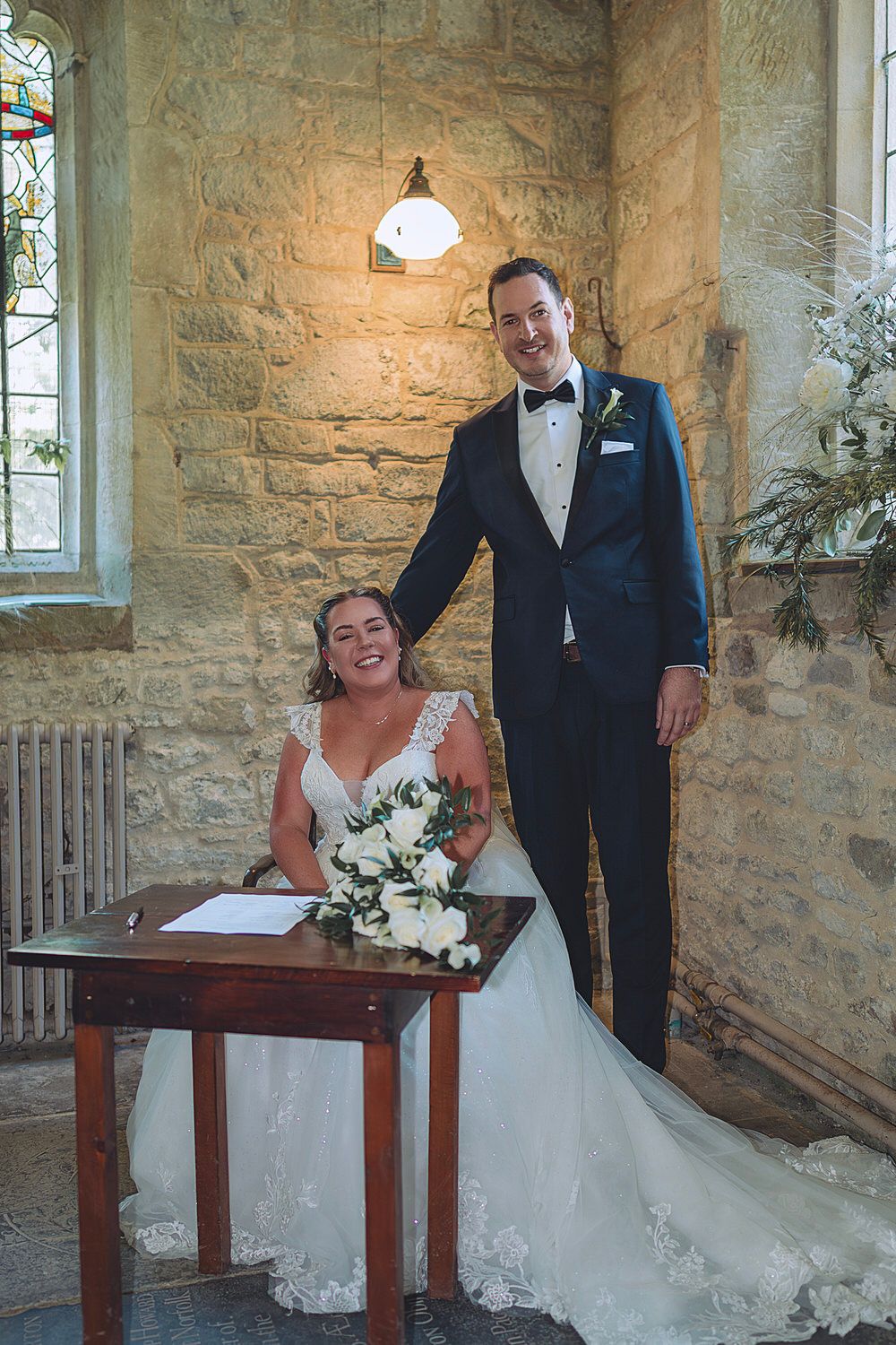 Bride and groom signing wedding register in stone church, bride in lace gown with white bouquet, groom in navy tuxedo.