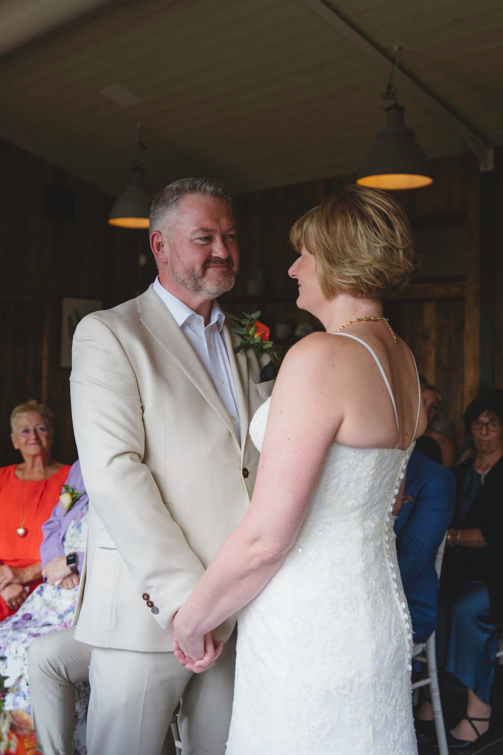 Bride and groom holding hands during wedding ceremony, groom in beige suit smiling at bride in white lace dress.