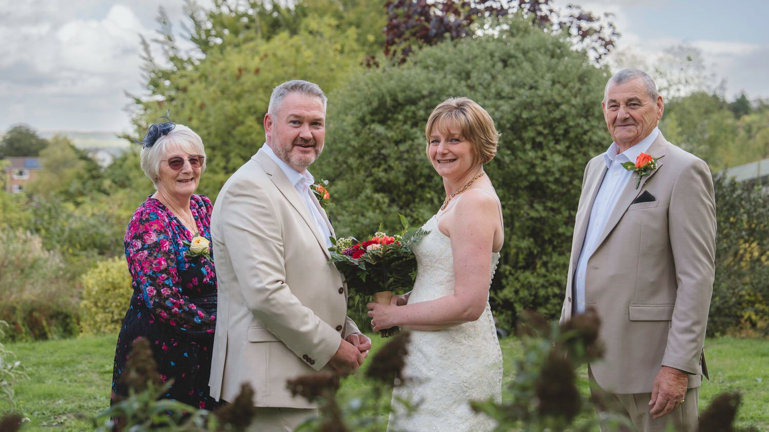 Wedding couple with family pose outdoors, bride holds bouquet, guests in formal attire on green lawn.