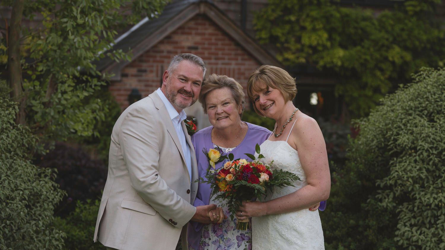 Wedding couple poses with smiling woman holding bouquet outdoors near brick building.