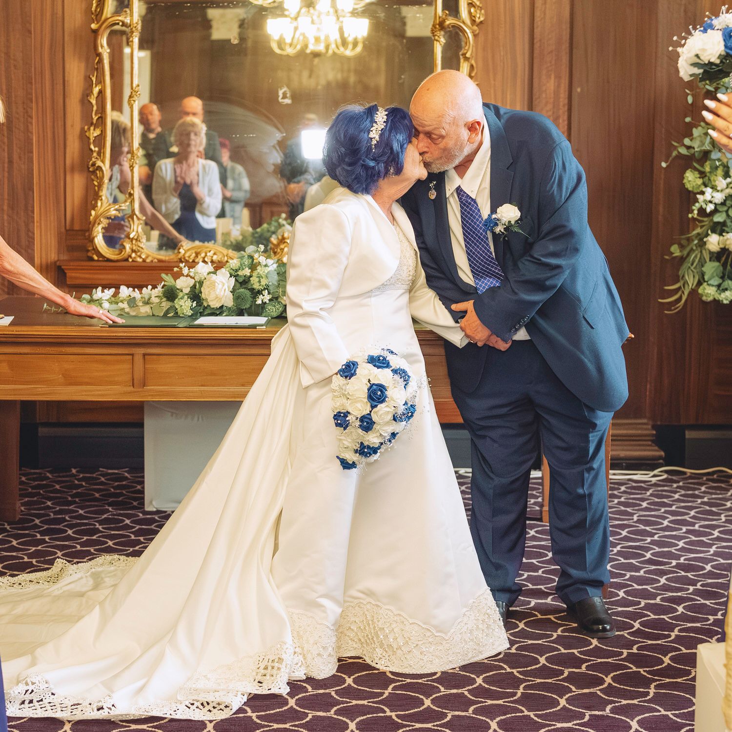 Bride and groom sharing a kiss at wedding ceremony, bride in white gown holding blue bouquet, groom in navy suit.
