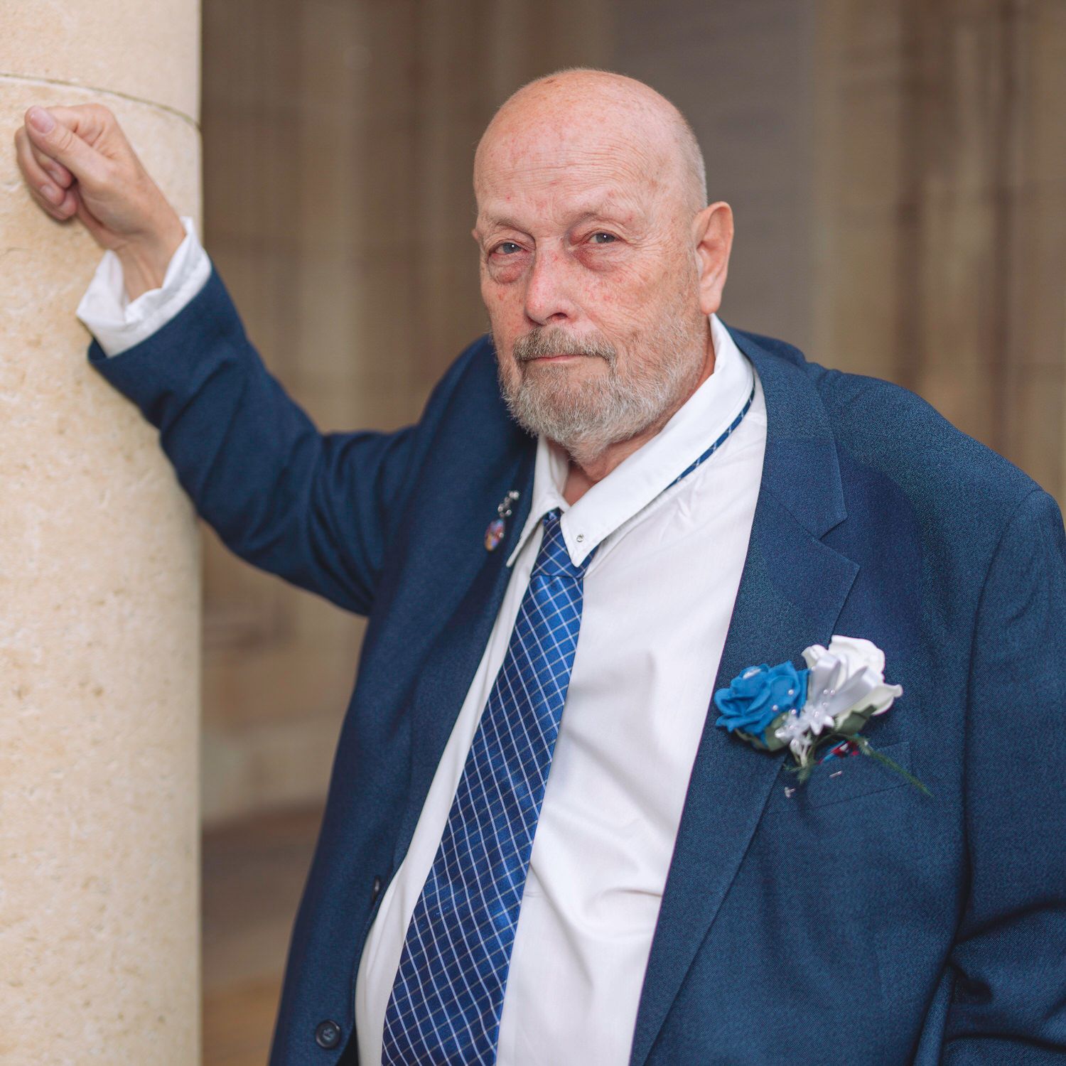 Elderly man in blue suit with boutonniere leaning against stone column.