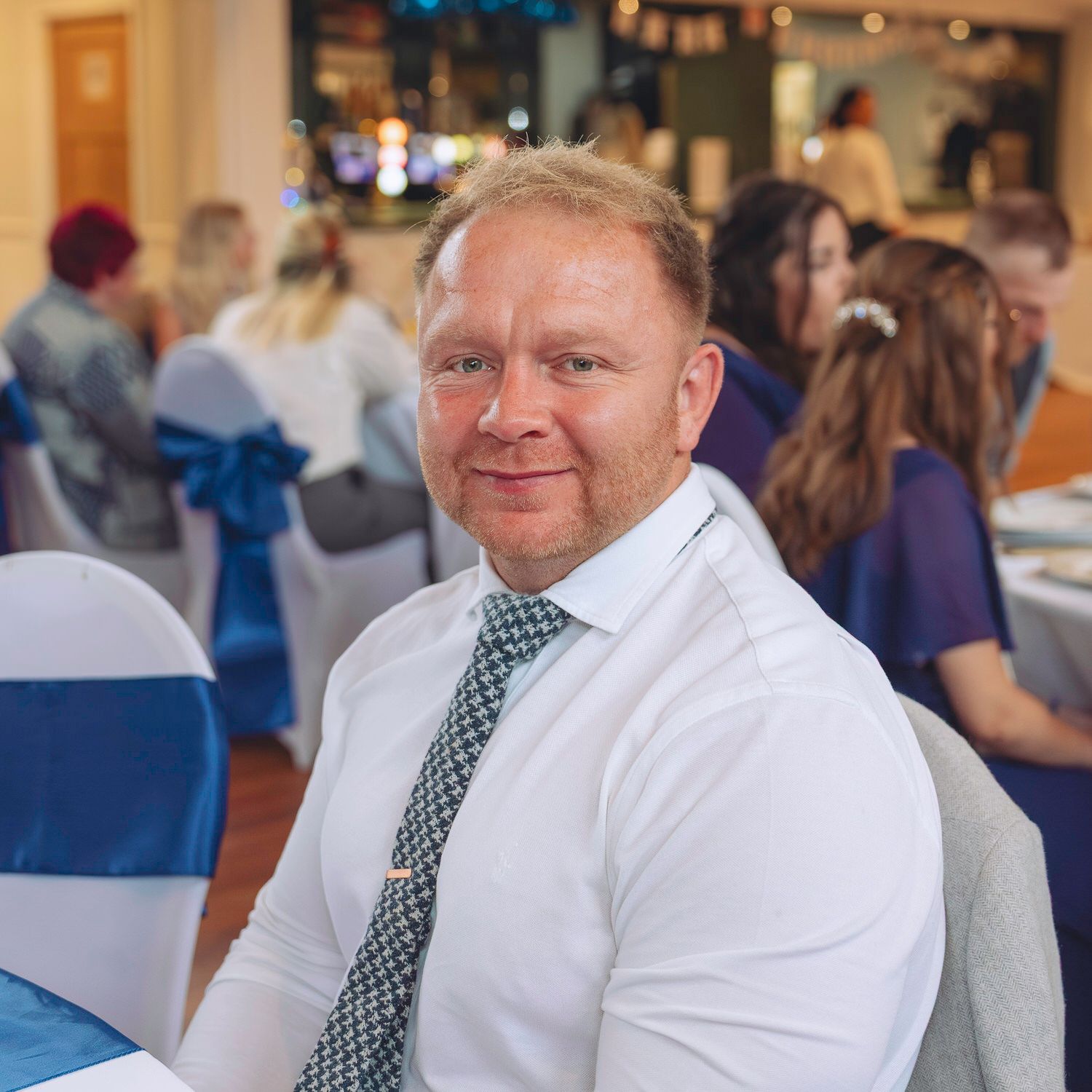 Smiling man in white shirt and tie seated at a decorated event venue with blue chair sashes.