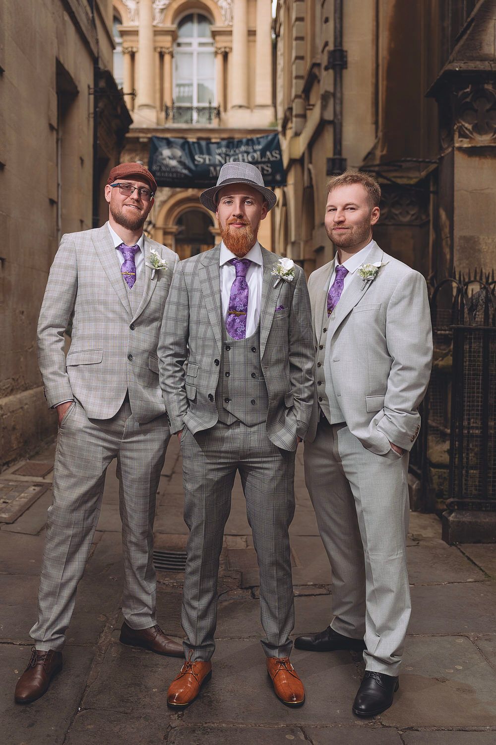 Three groomsmen in gray suits and purple ties pose in a historic alley, groom in center wearing a hat and vest.