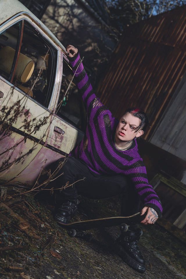 Person in purple striped sweater crouching with skateboard near abandoned car in urban decay setting.