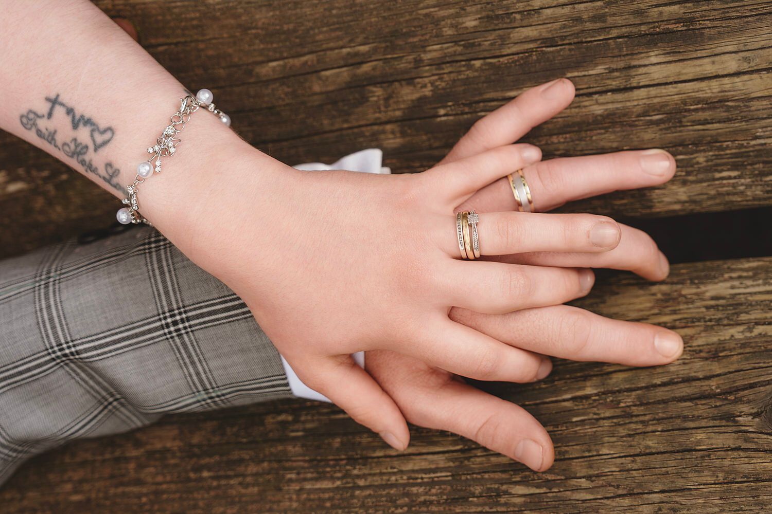 Two hands resting on wood, one with a cross tattoo and charm bracelet, the other wearing elegant stacked gold rings.