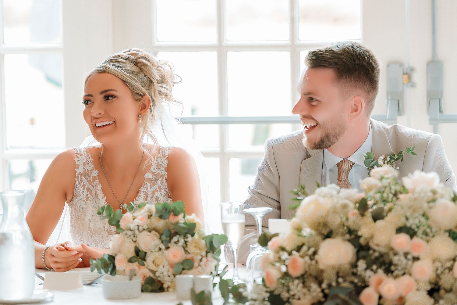 Happy bride and groom laughing at wedding reception table with elegant floral arrangements.