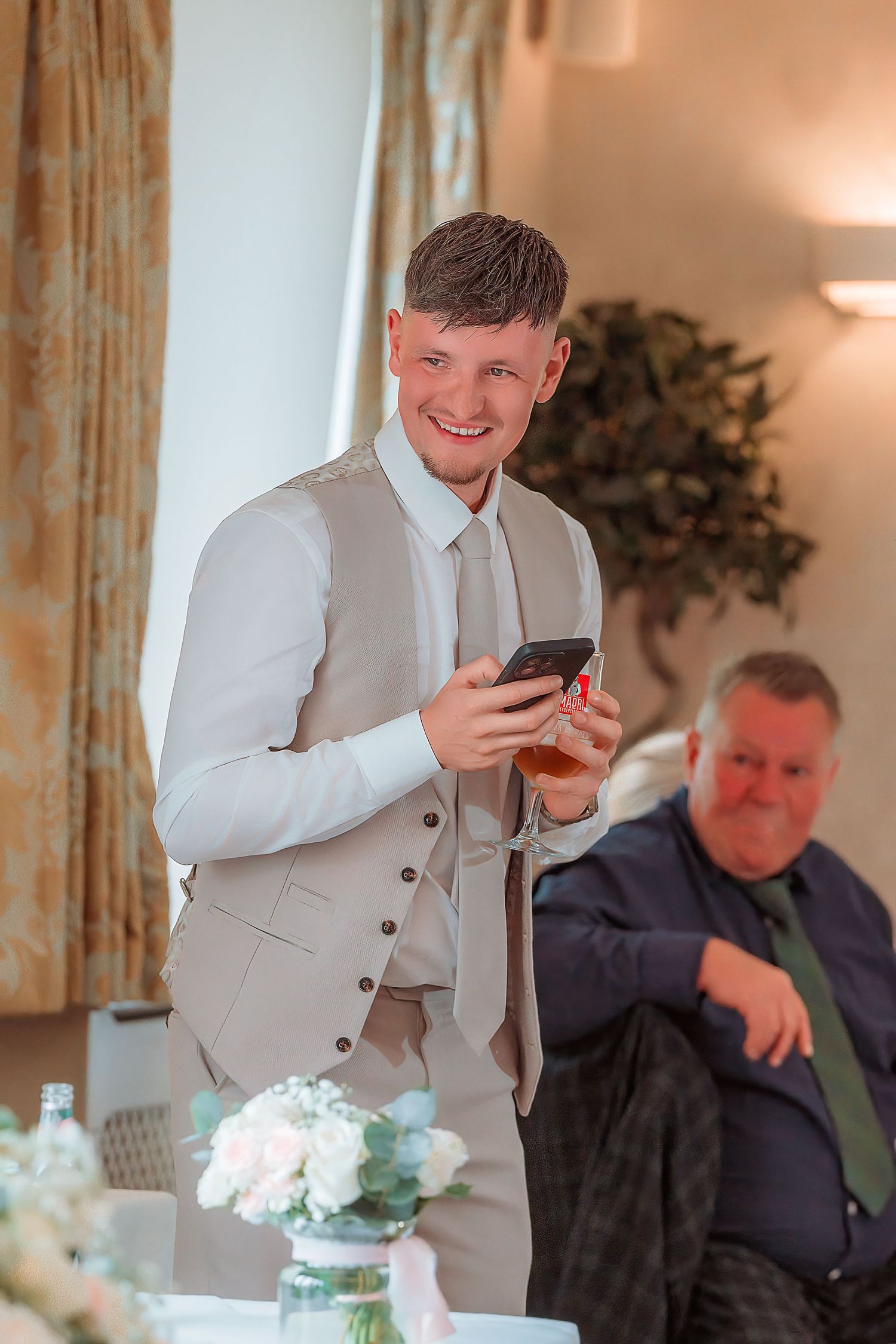 Smiling young man in beige suit vest holding phone and drink at elegant wedding reception.