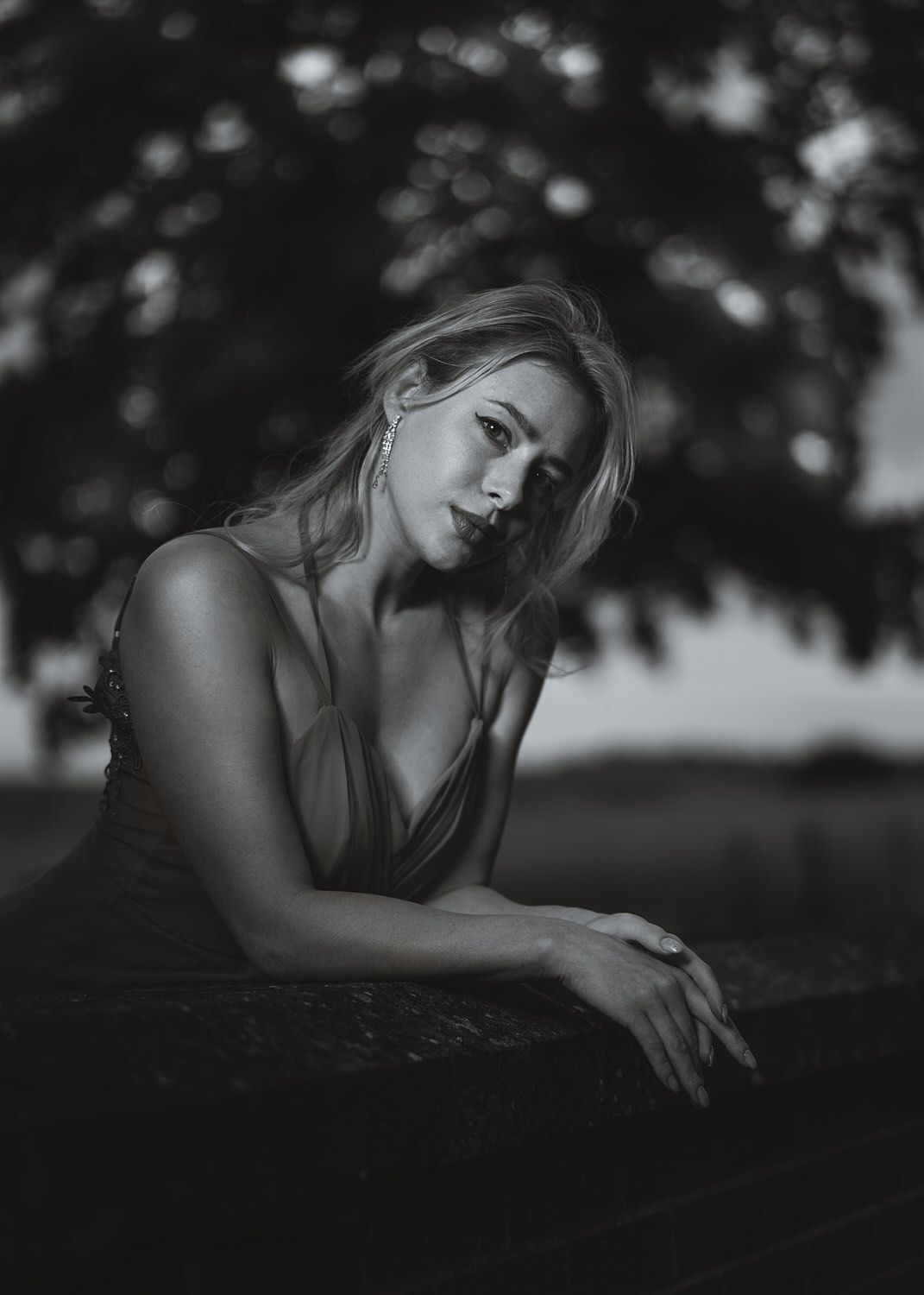 Black and white portrait of a blonde woman leaning on a surface outdoors with blurred trees in the background.