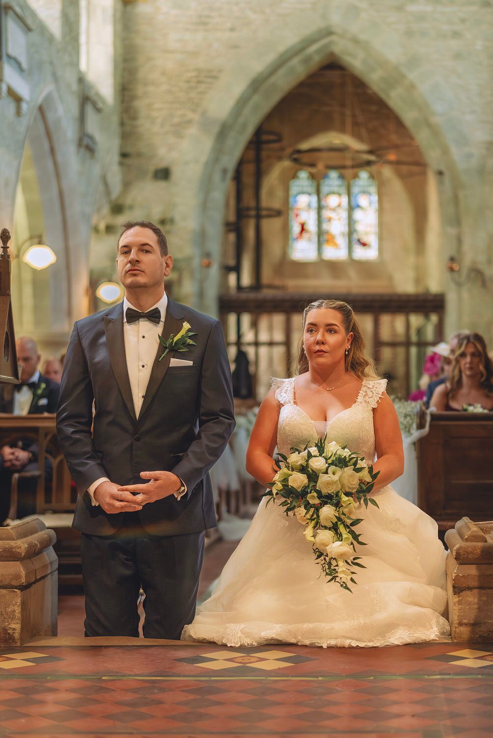 Bride holding white bouquet and groom in navy tuxedo stand at church altar during wedding ceremony.