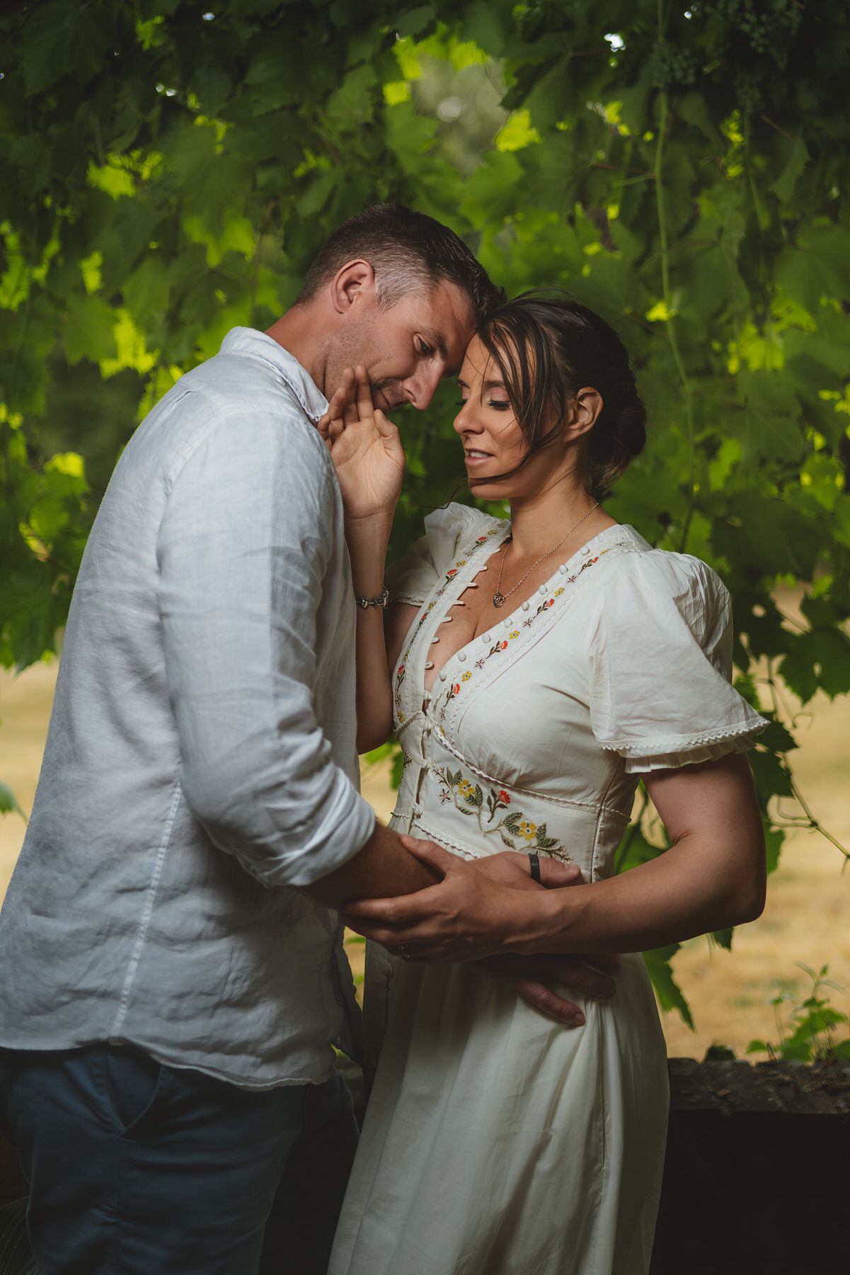 Couple share intimate romantic moment together beneath green leafy canopy in warm summer light.