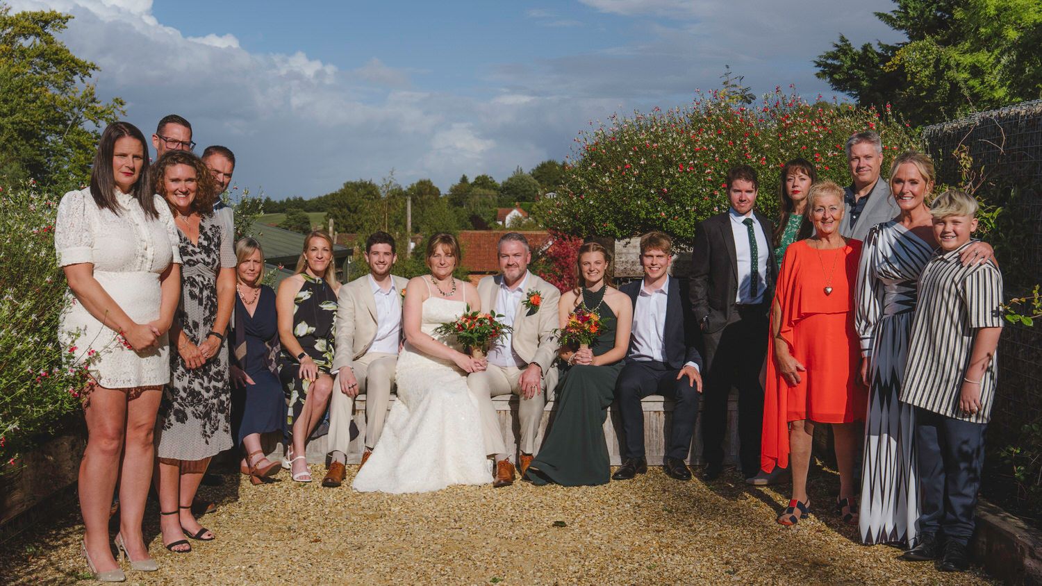 Wedding group photo with bride and groom surrounded by family and friends outdoors on a sunny day.
