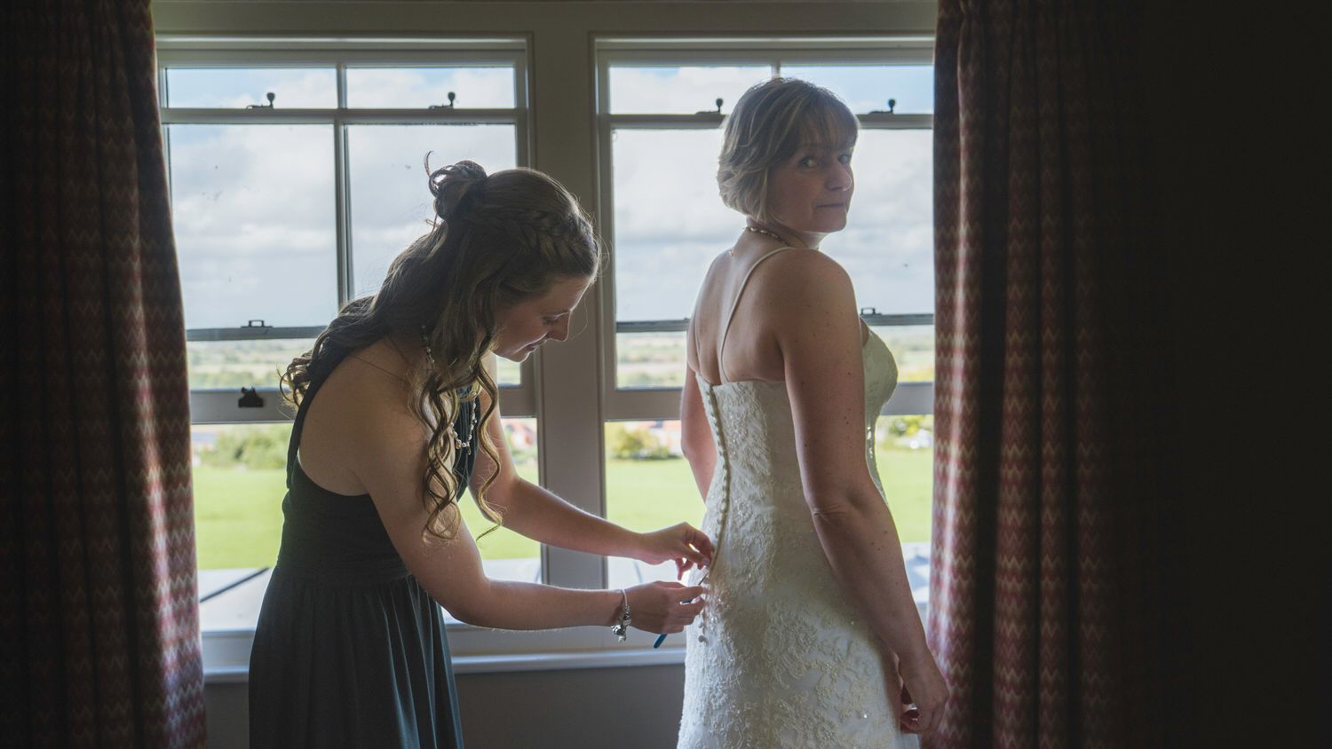 Bridesmaid helping bride fasten her lace wedding dress by a bright window on the wedding day.