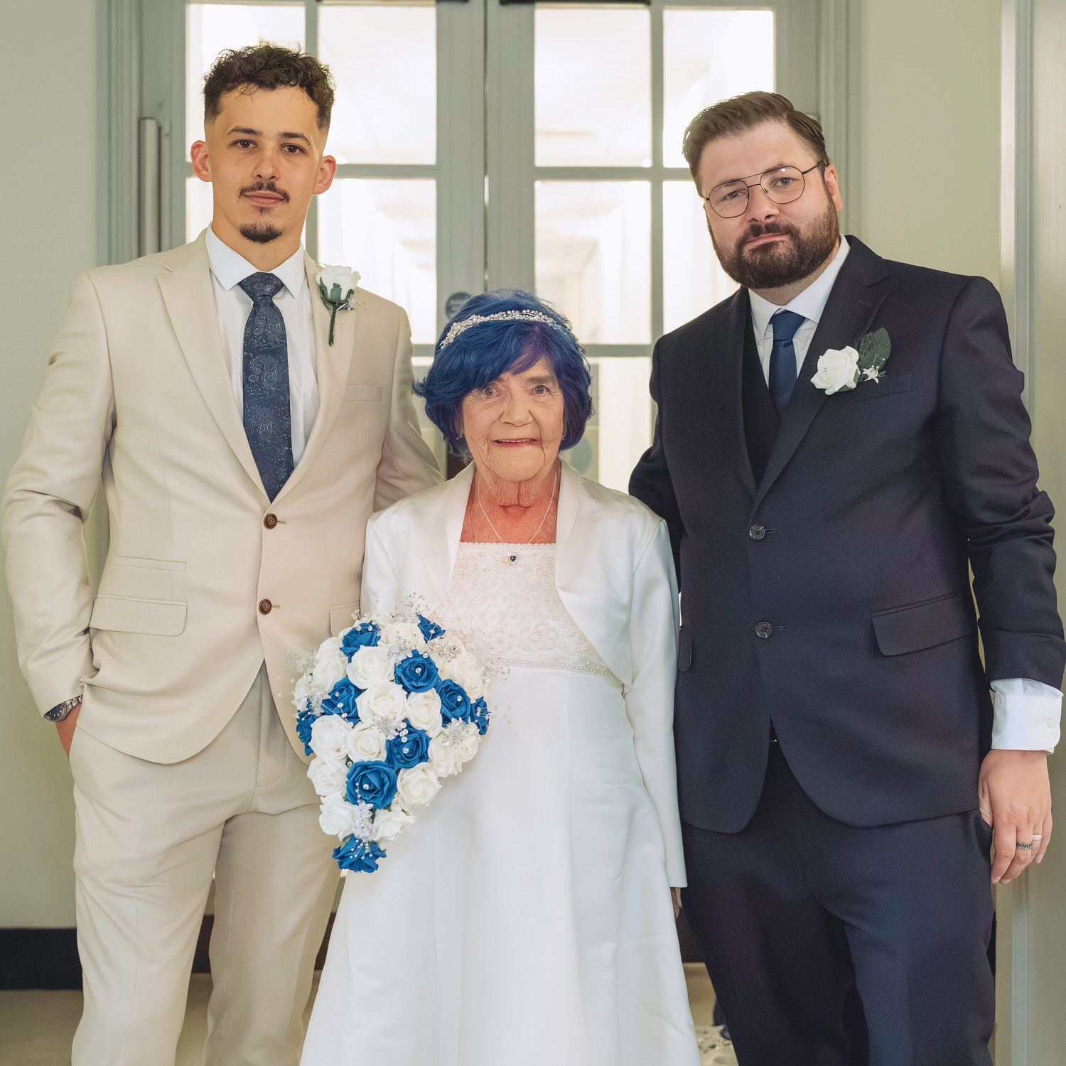Elderly bride with blue hair holds a blue and white bouquet flanked by two suited groomsmen at a wedding.