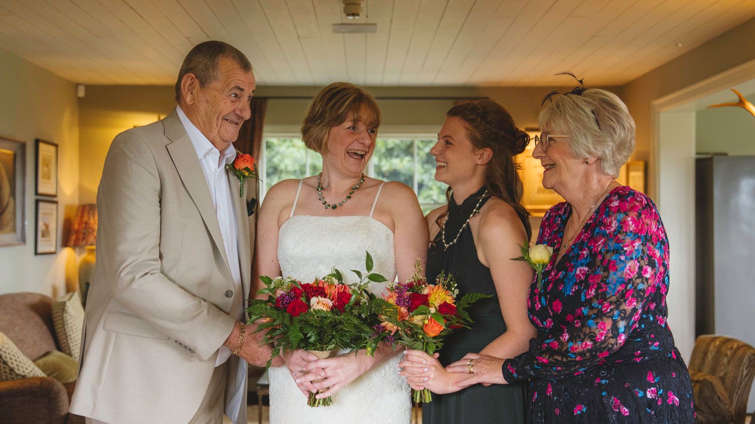 Bride in white dress holds colorful bouquet, smiling with three guests in a warmly lit indoor wedding setting.