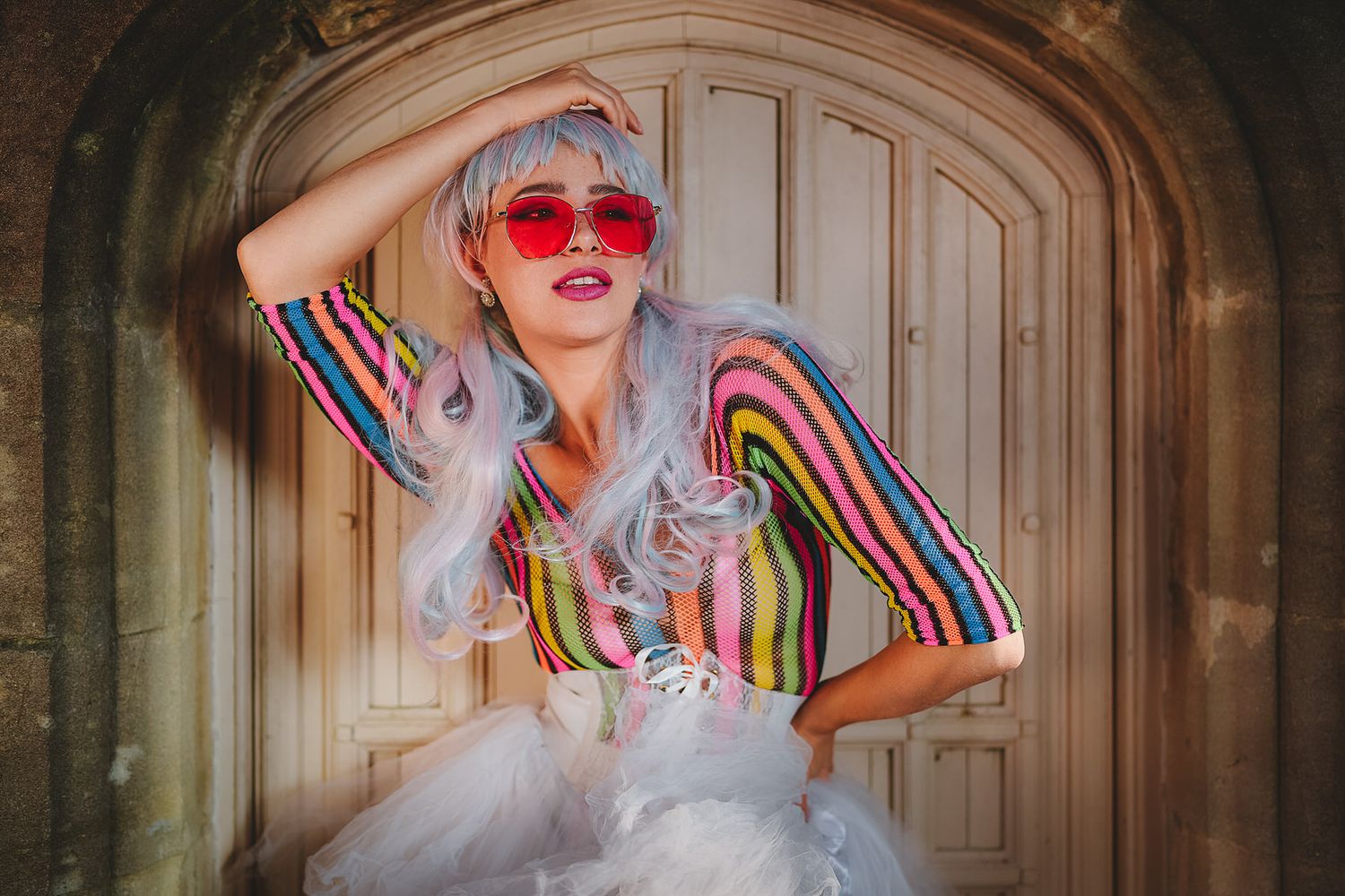 Woman in rainbow striped top, silver wig, and red heart sunglasses posing before ornate wooden door.