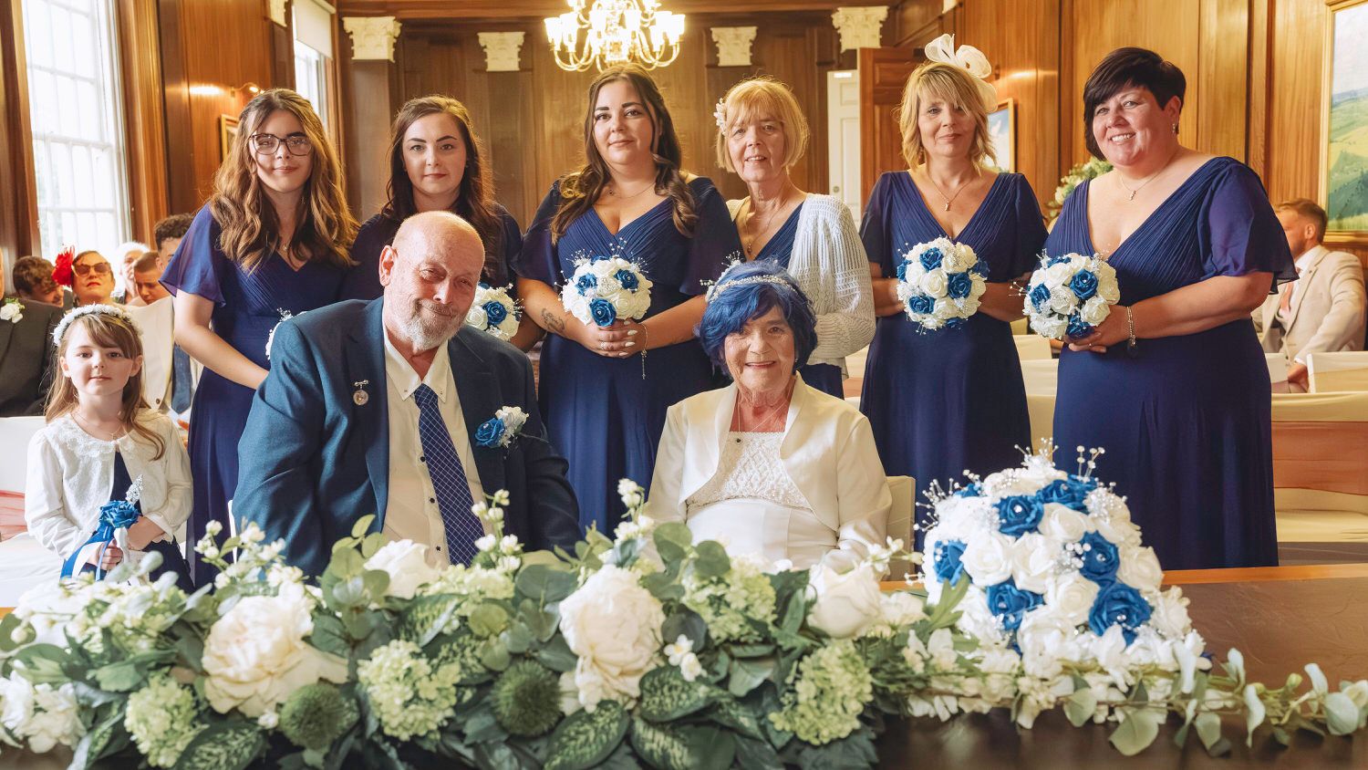 Elderly couple seated at wedding table with bridesmaids in navy blue dresses holding teal bouquets in elegant venue.
