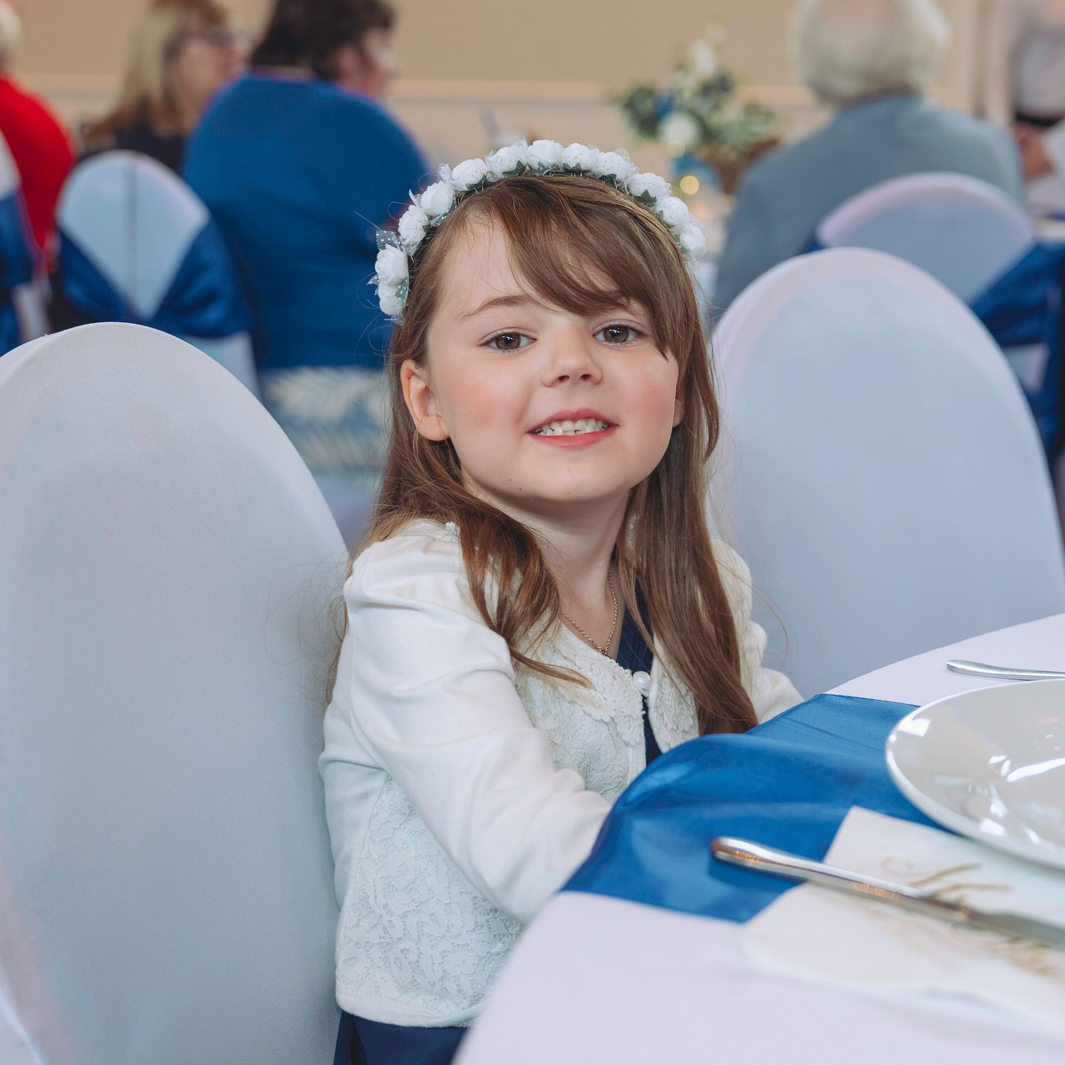 Young girl in white floral headband smiles at wedding reception table with blue satin runner and formal place settings.