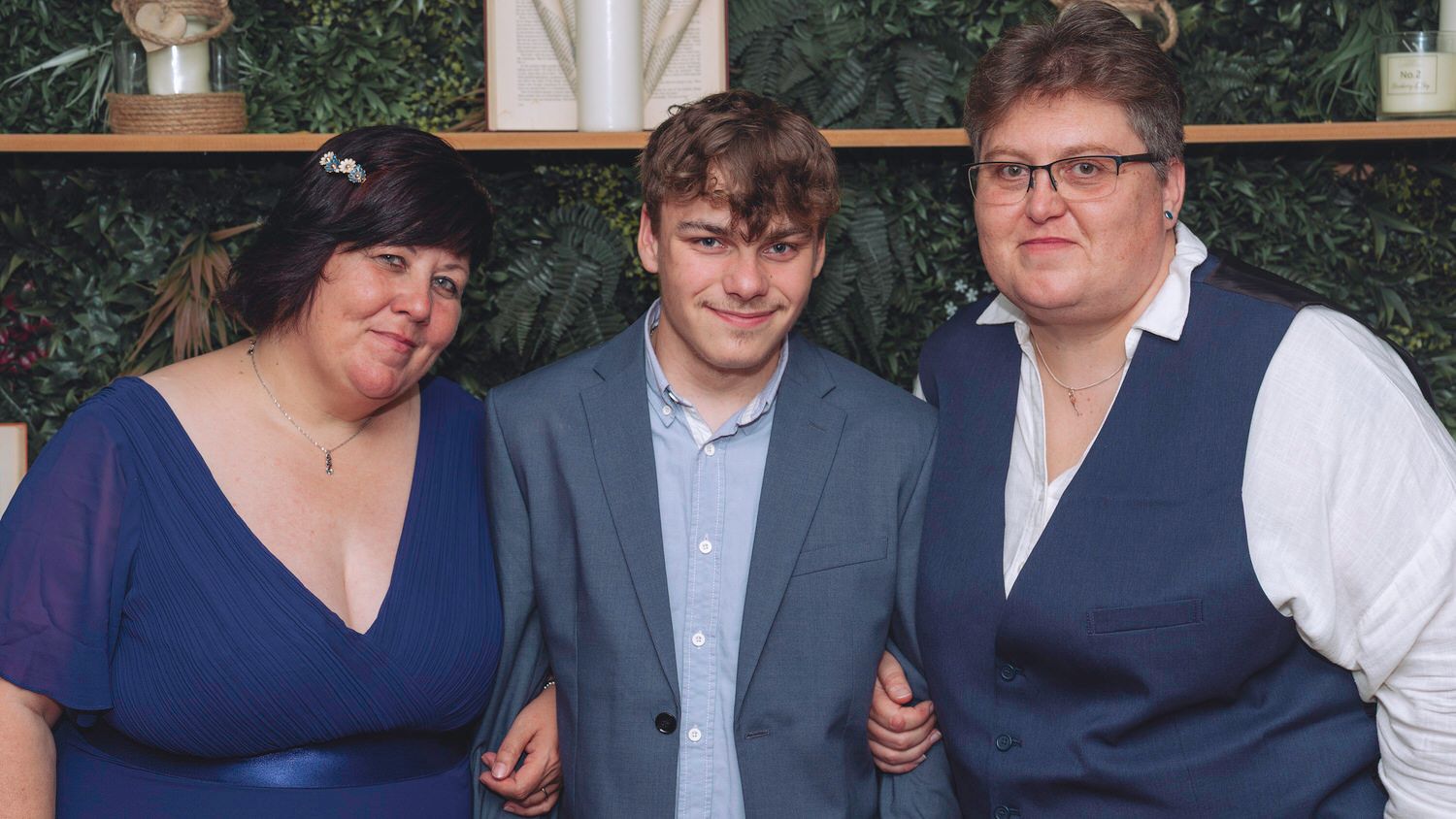 Three people posing together at a formal event in front of a green floral wall backdrop.