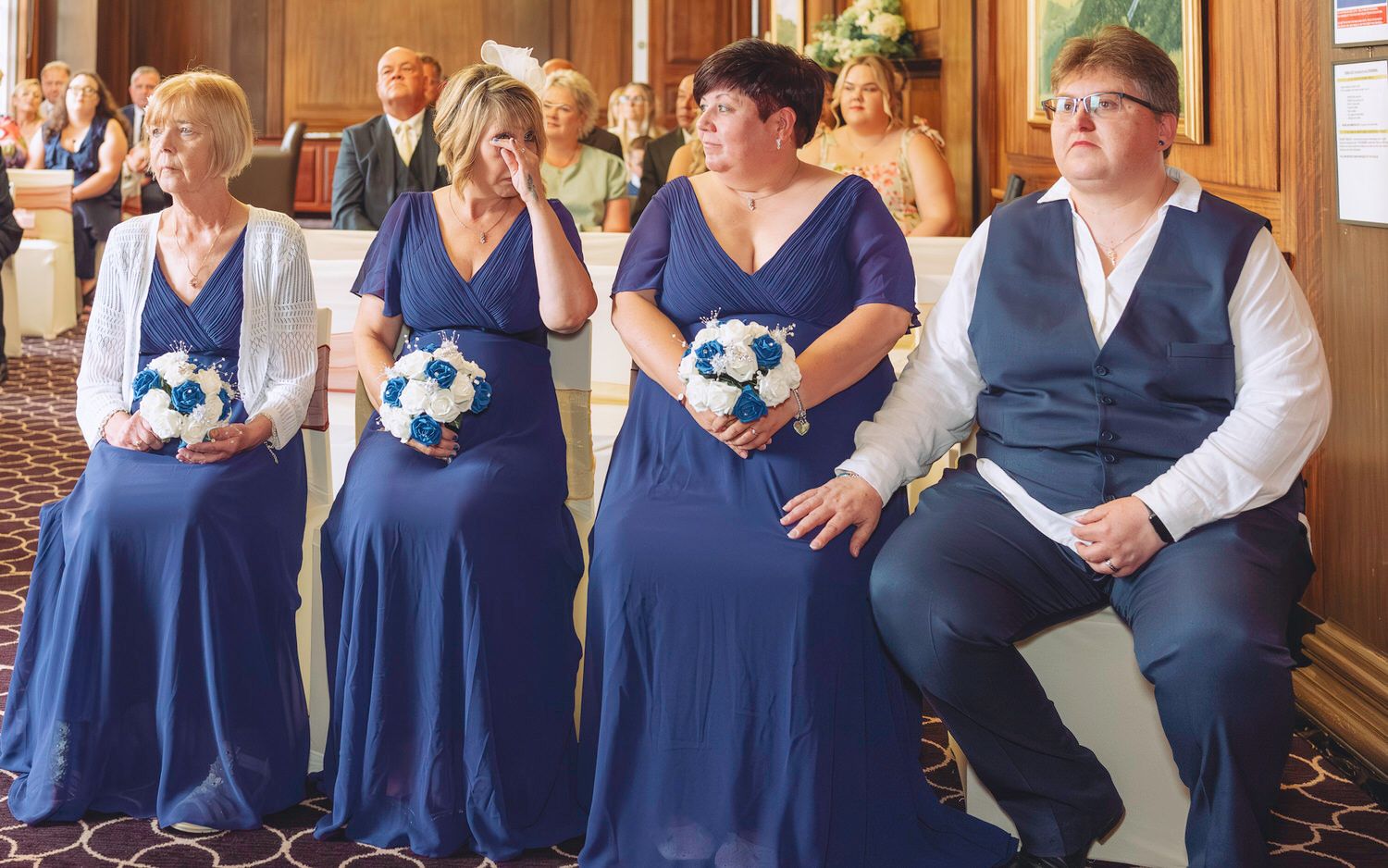 Four bridesmaids in navy blue dresses holding white bouquets stand together during a wedding ceremony indoors.