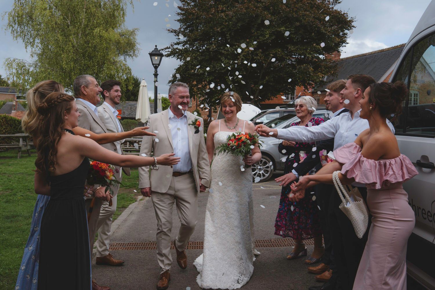 Bride and groom walk through confetti shower surrounded by smiling guests outside a charming countryside wedding venue.