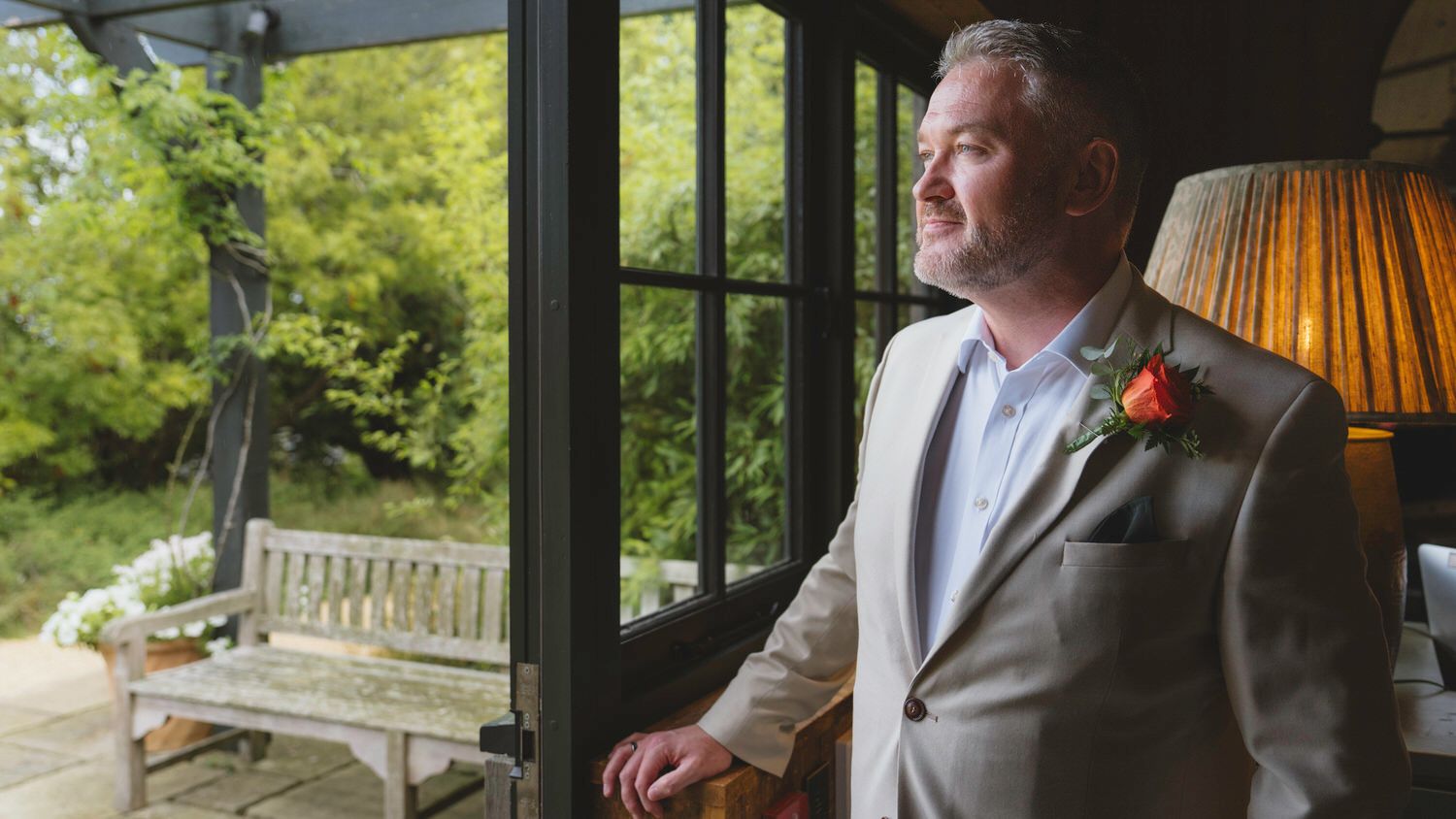 Groom in gray suit with boutonniere gazing out glass doors at garden with wooden bench.
