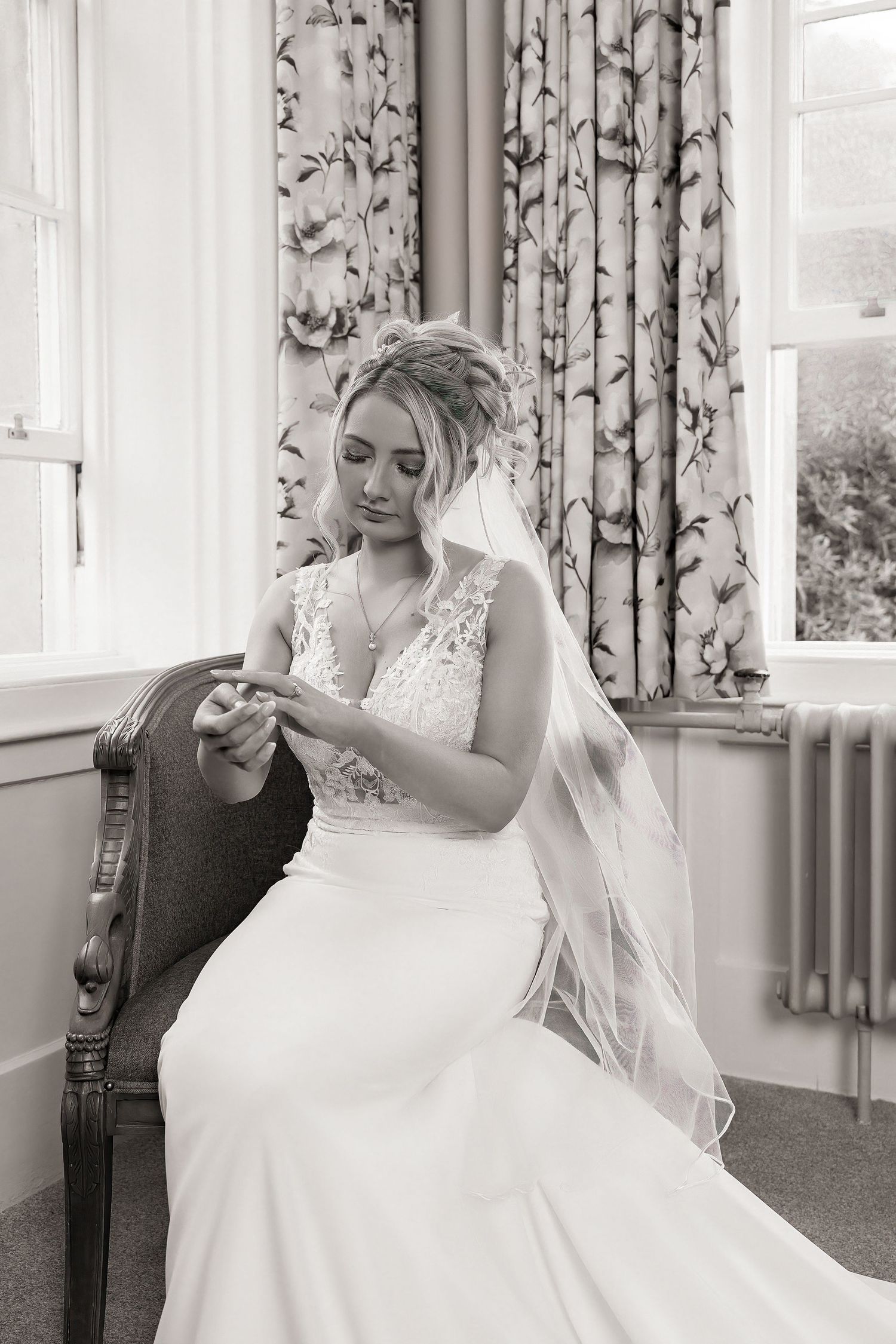Black and white photo of a bride in a lace wedding dress and veil, seated in a chair by a window.