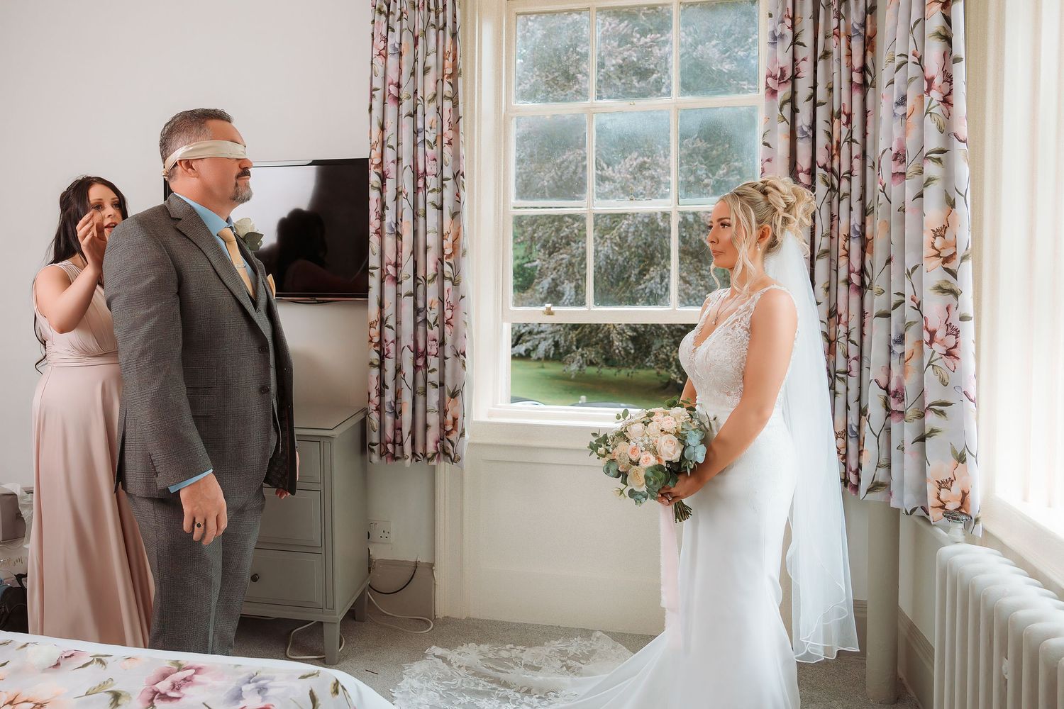 Bride in white gown holds bouquet as emotional father sees her for first time on wedding day in bright bedroom.