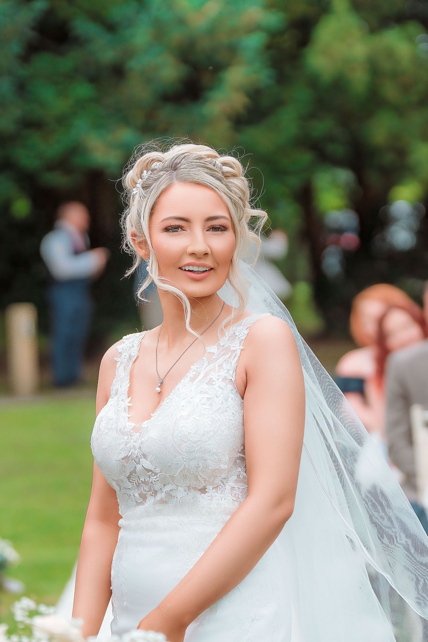 Smiling bride in lace wedding dress and veil poses outdoors with green trees in background.