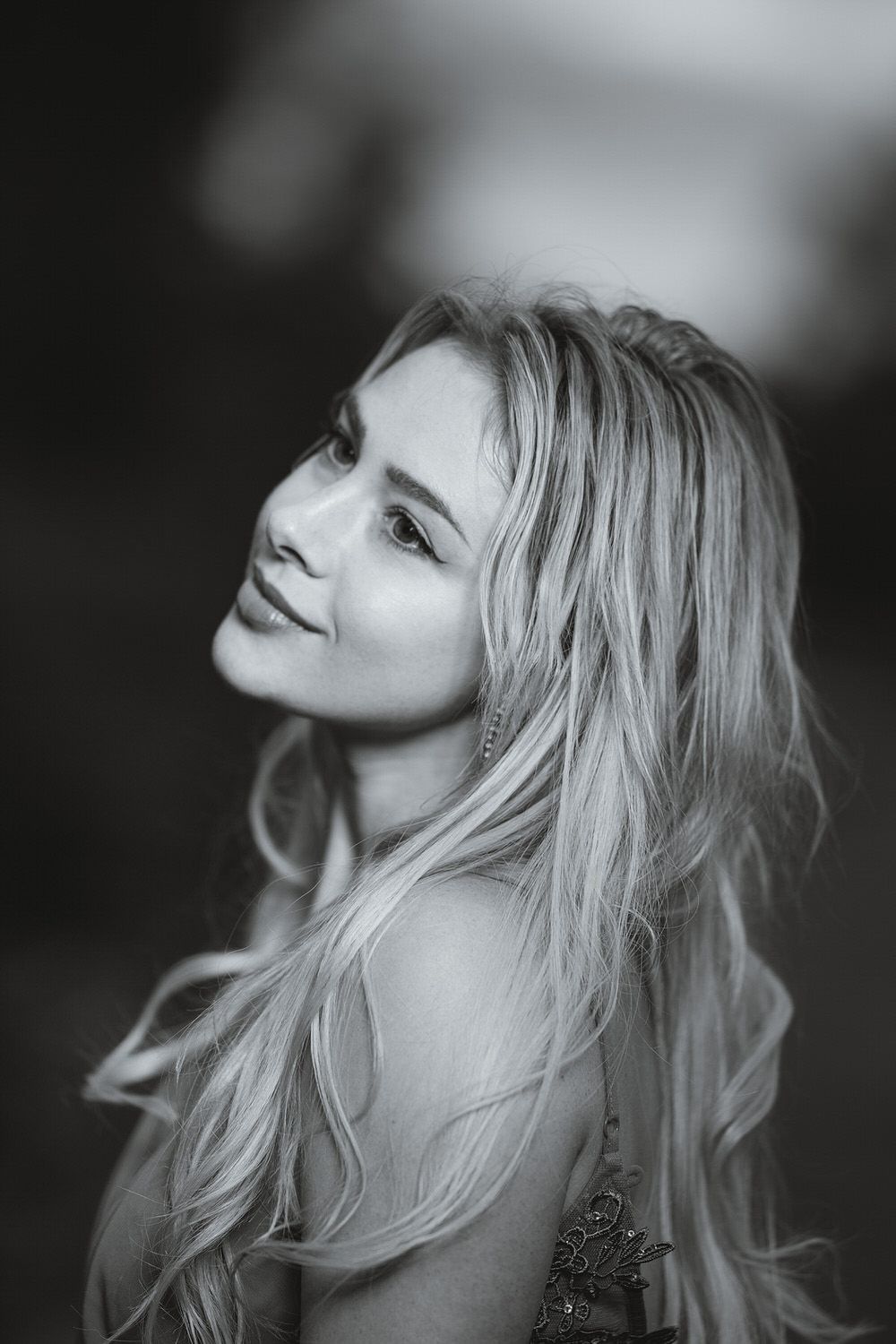 Black and white portrait of a smiling blonde woman with long wavy hair, gazing upward against a blurred background.