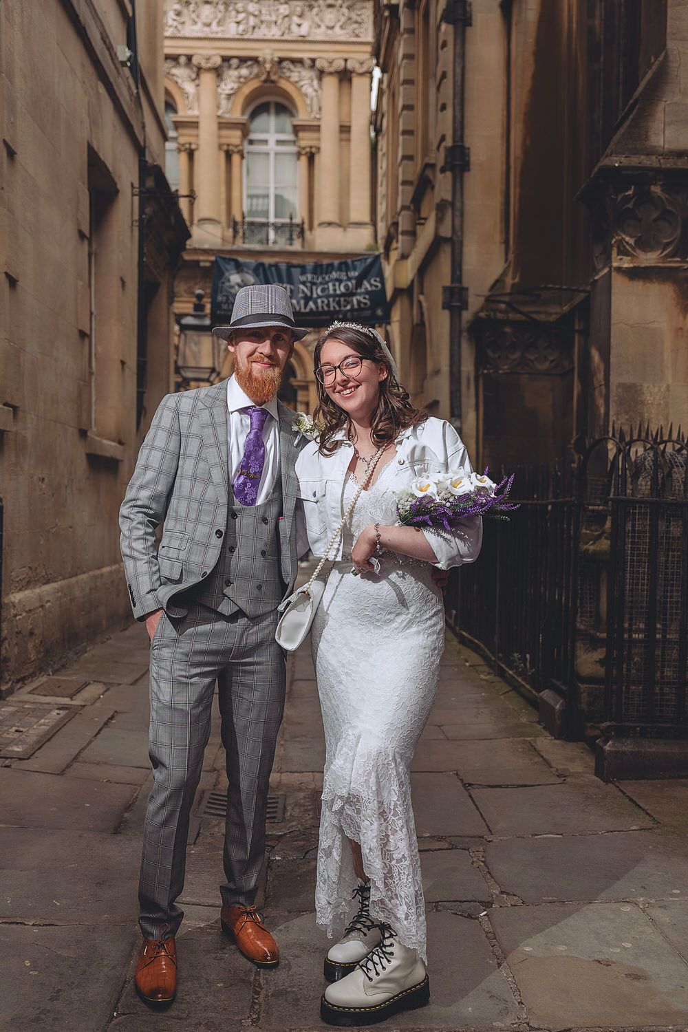 Bride in lace dress and groom in grey plaid suit pose together in a historic cobblestone alley near Nicholas Markets.
