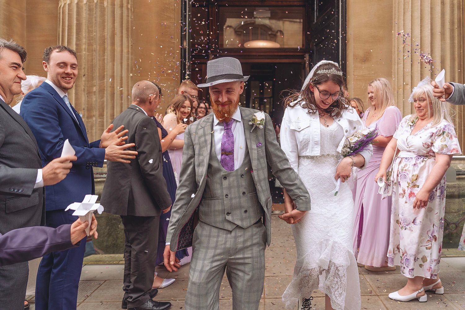 Bride and groom exit church amid confetti shower, surrounded by smiling wedding guests on sunny day.