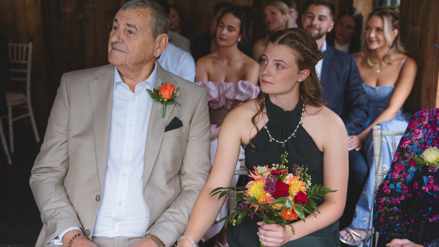 Man and woman in formal attire seated at a wedding ceremony, woman holding a colorful bouquet of flowers.