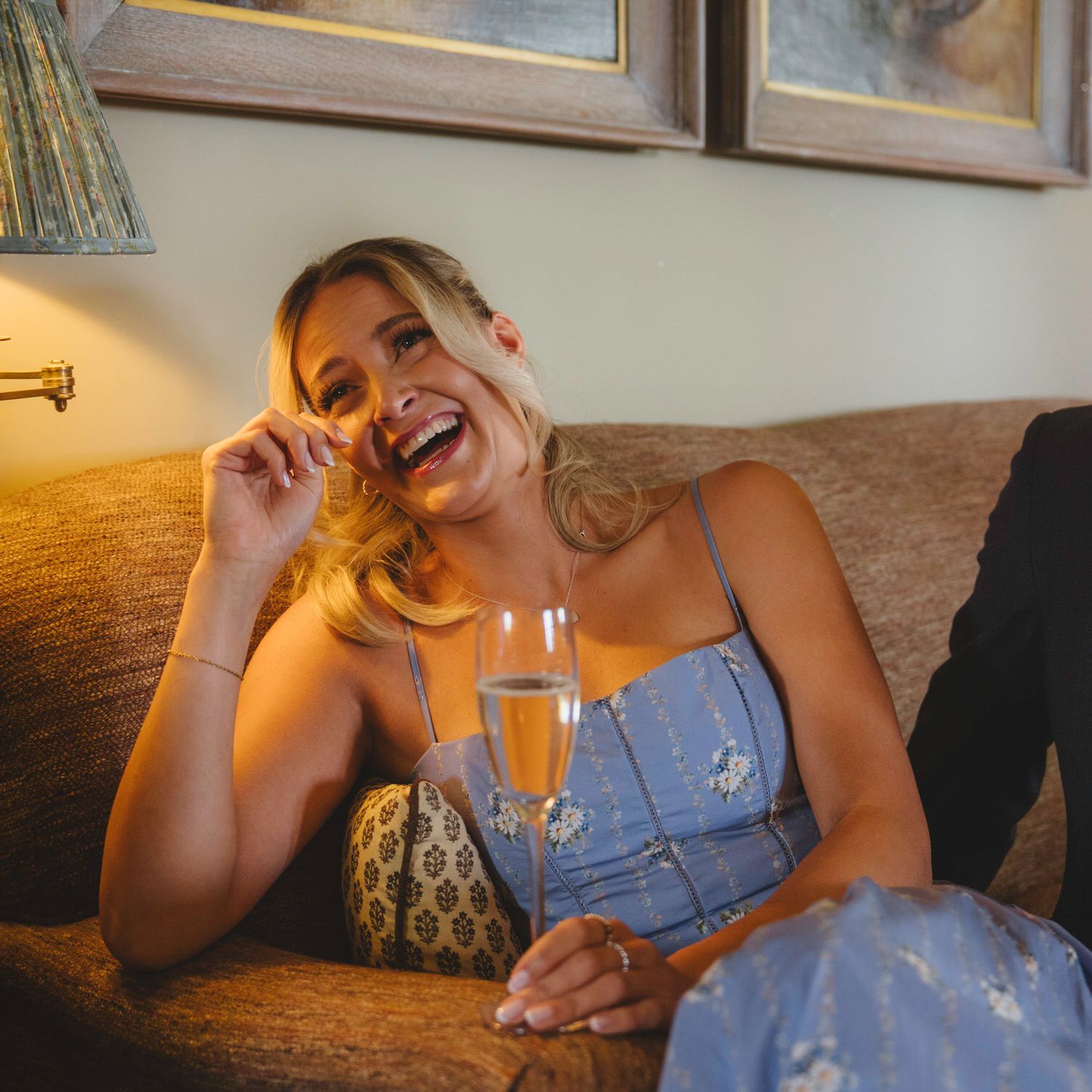 Woman in blue dress laughing on sofa holding champagne flute at elegant indoor gathering.