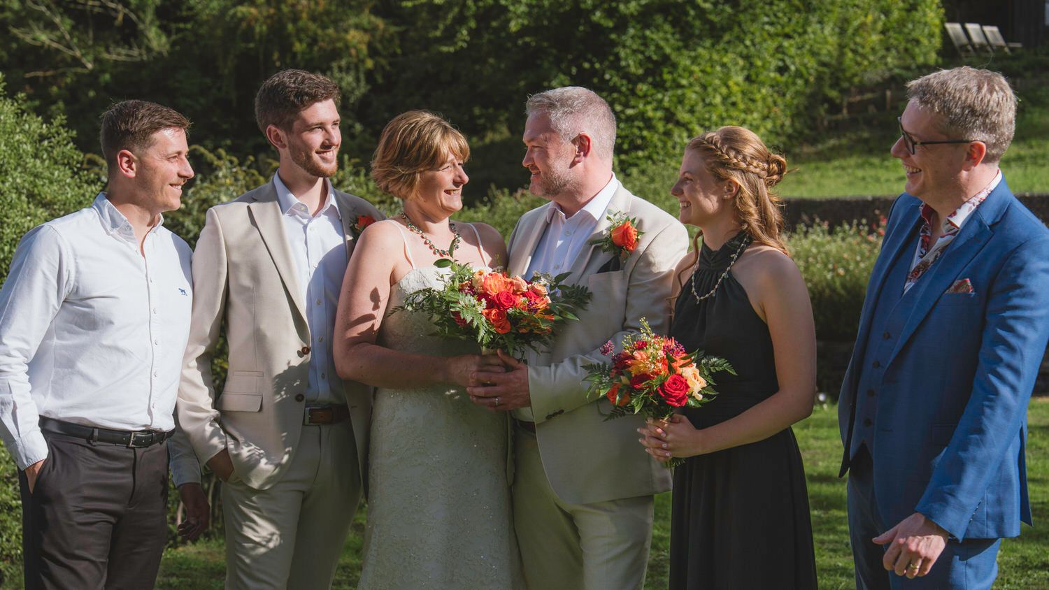 Wedding party of six people smiling outdoors, bride holding colorful bouquet in lush green garden setting.