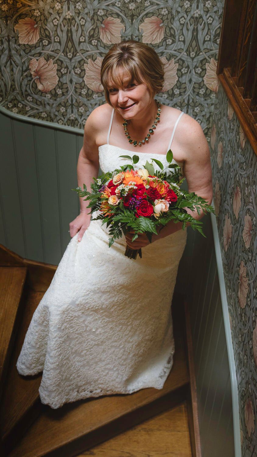 Bride in white lace gown holding red and orange bouquet descending wooden staircase with floral wallpaper.