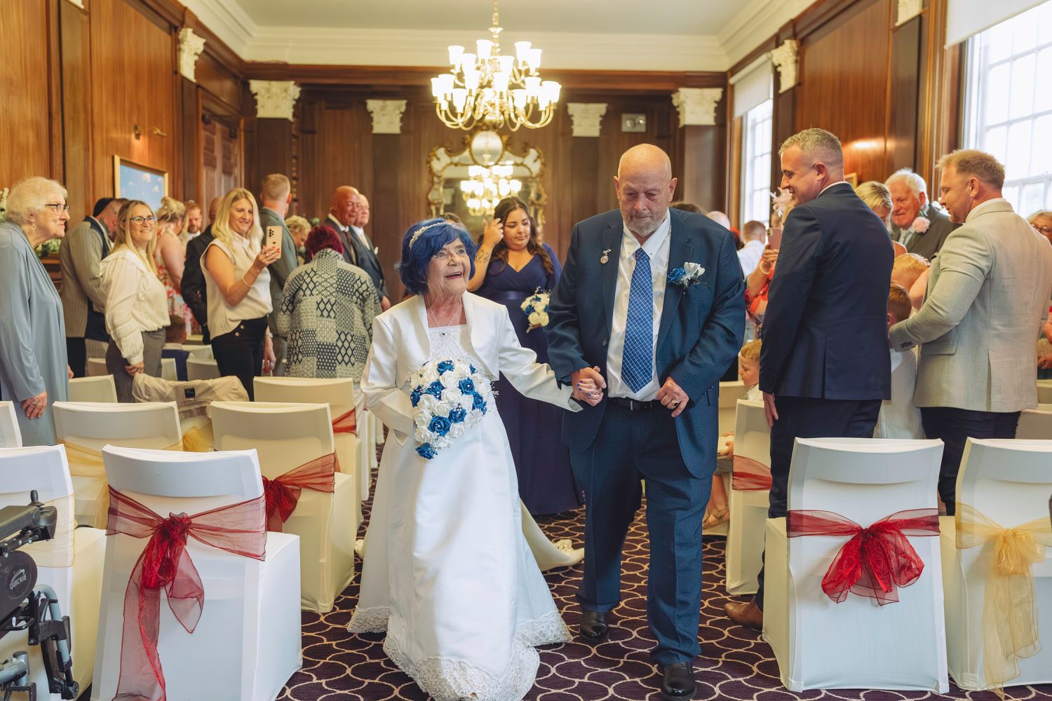 Elderly couple walks down the aisle at a wedding ceremony in an elegant room with chandelier and guests.