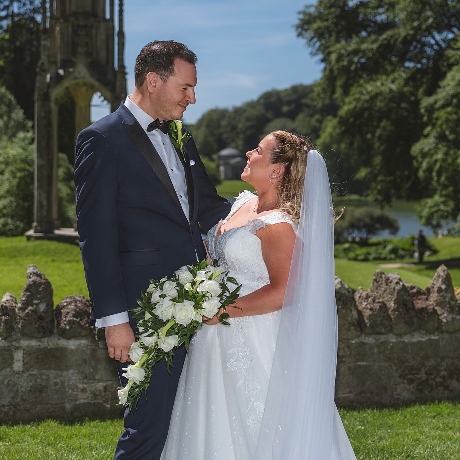 Wedding couple share romantic gaze near ancient stone ruins with lake and trees in background.
