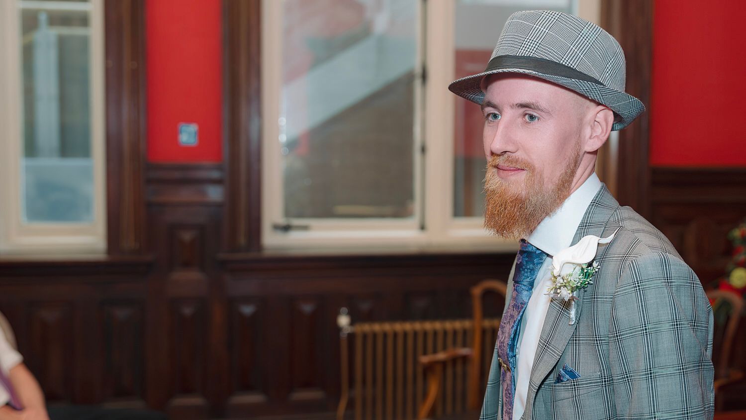Bearded ginger man in plaid suit and grey fedora wearing boutonniere at formal event.