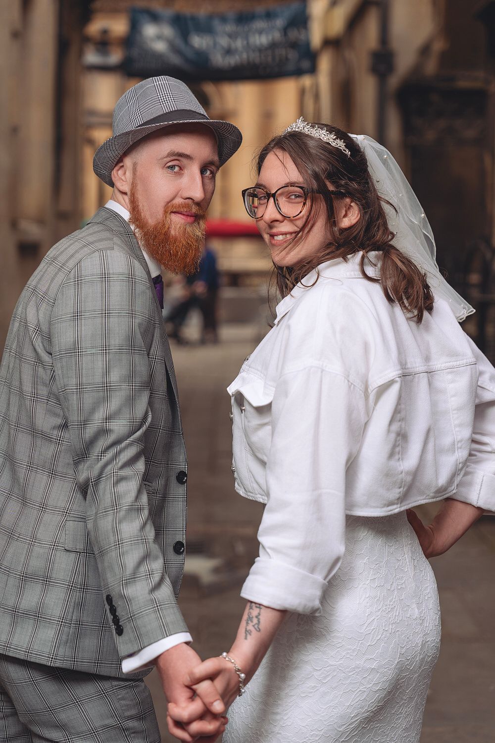 Smiling newlywed couple holding hands in an alley, groom in gray suit and hat, bride in white dress and veil with glasses.