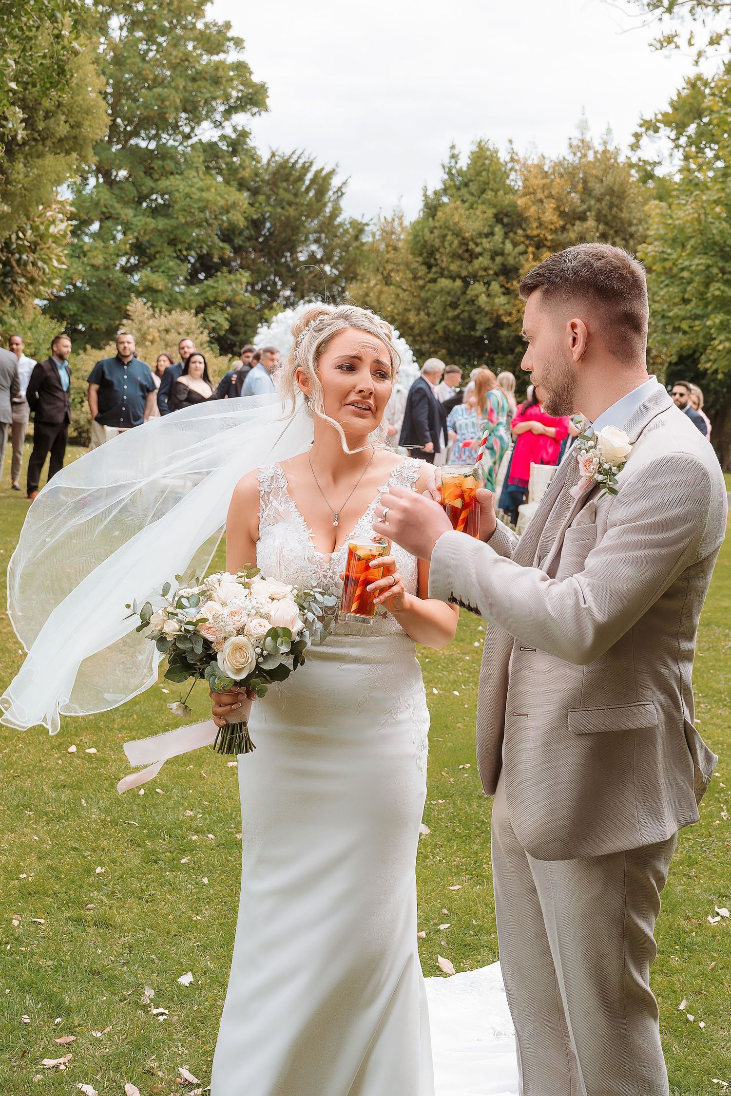 Bride and groom toasting with drinks outdoors at garden wedding ceremony, bride holding white rose bouquet with veil blowing.