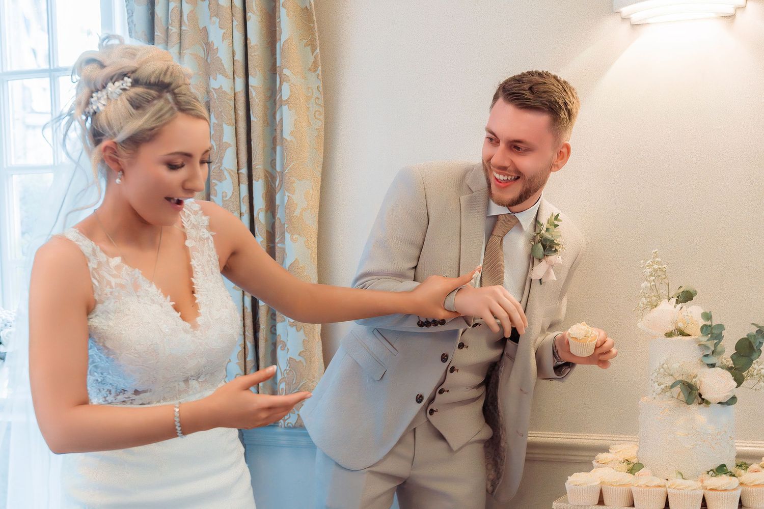 Bride and groom laughing while cutting their white wedding cake at an elegant indoor wedding reception.