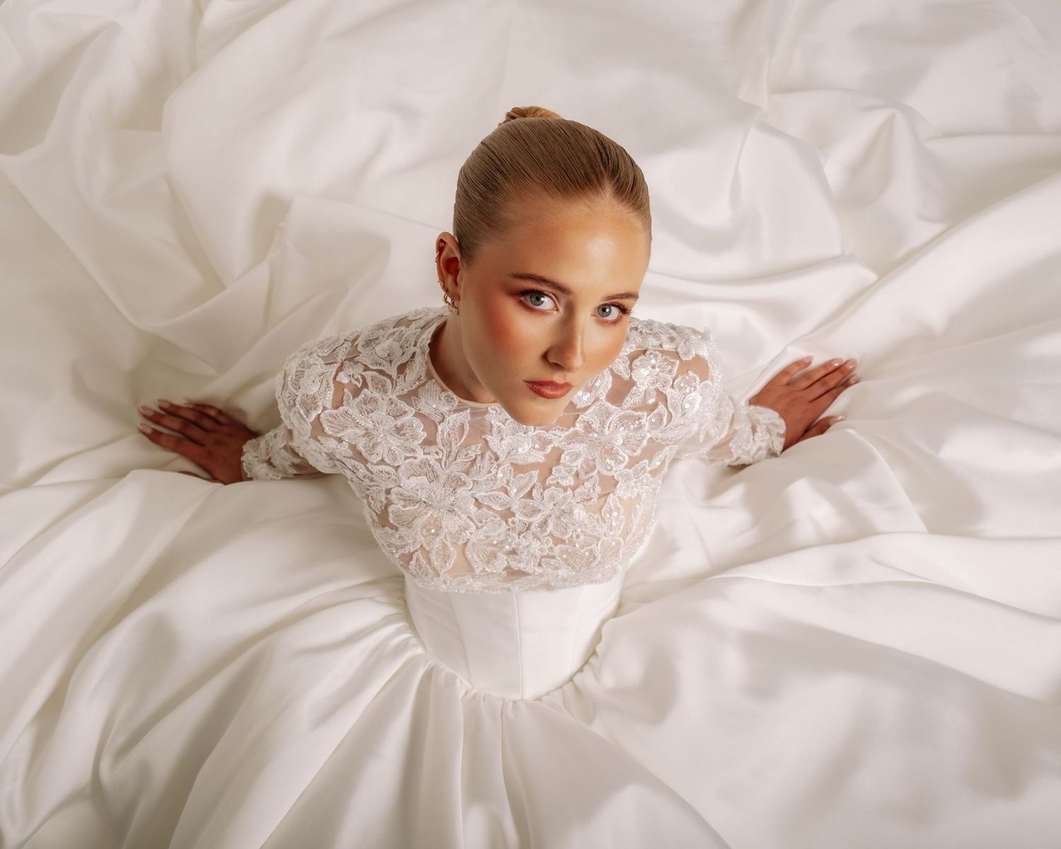 Bride in elegant white lace long-sleeve wedding gown sitting gracefully surrounded by flowing white fabric.
