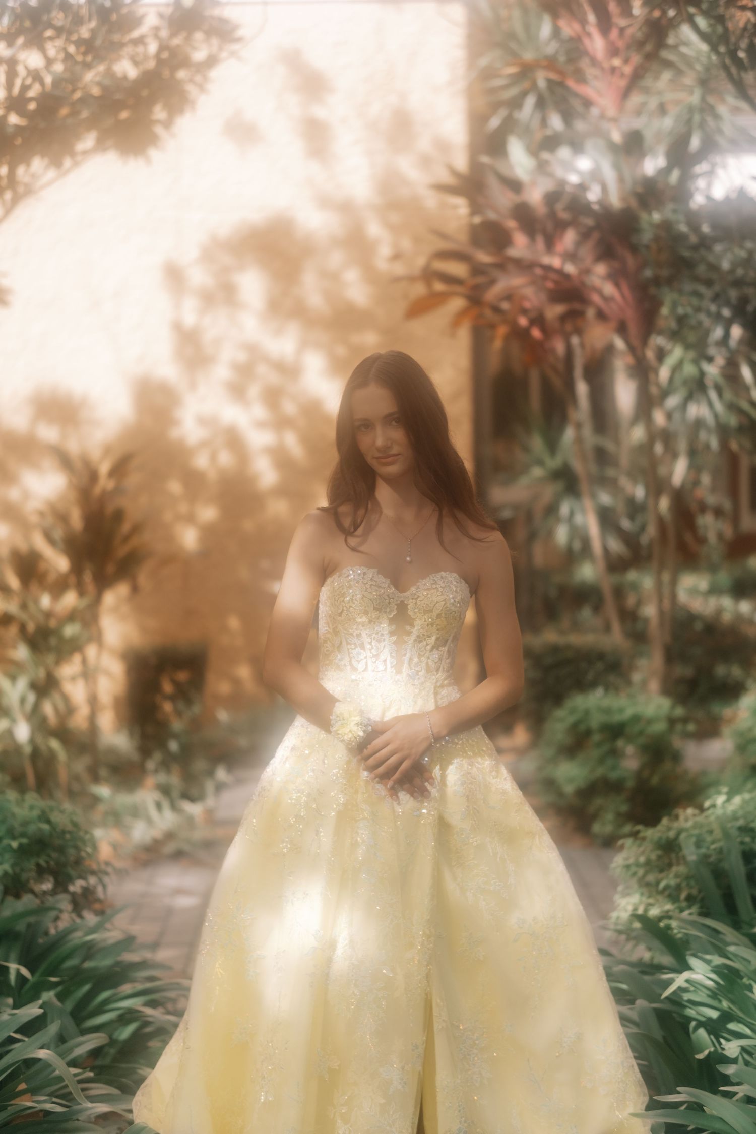 Woman in yellow strapless ball gown standing in lush garden with soft golden light.