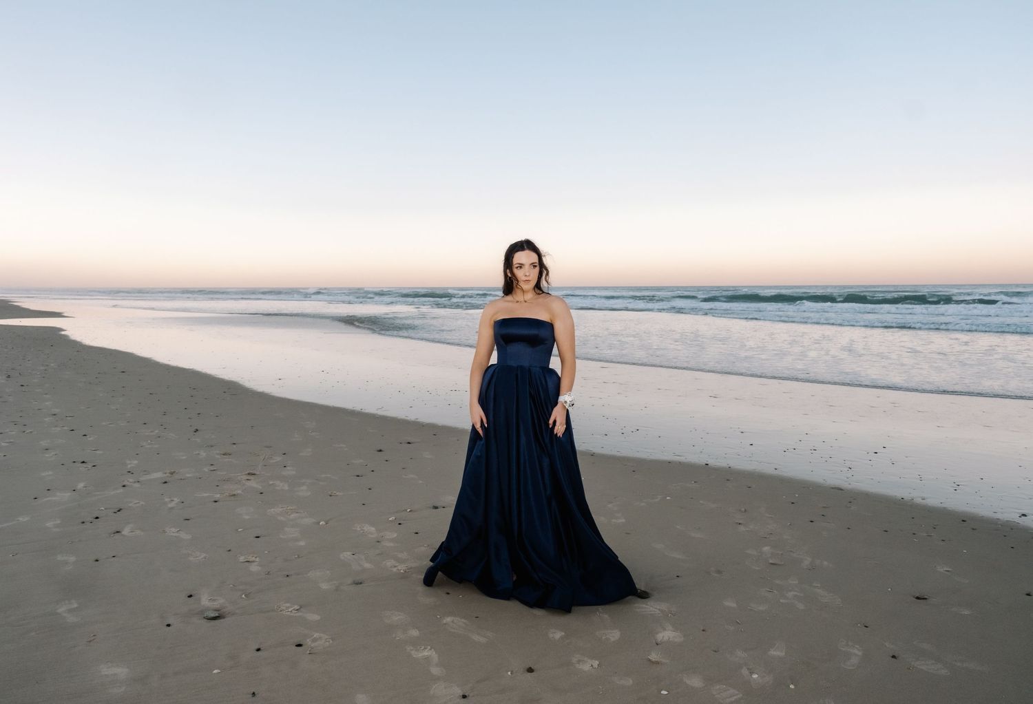 Woman in a strapless navy blue ballgown standing on a beach at sunset with city skyline.