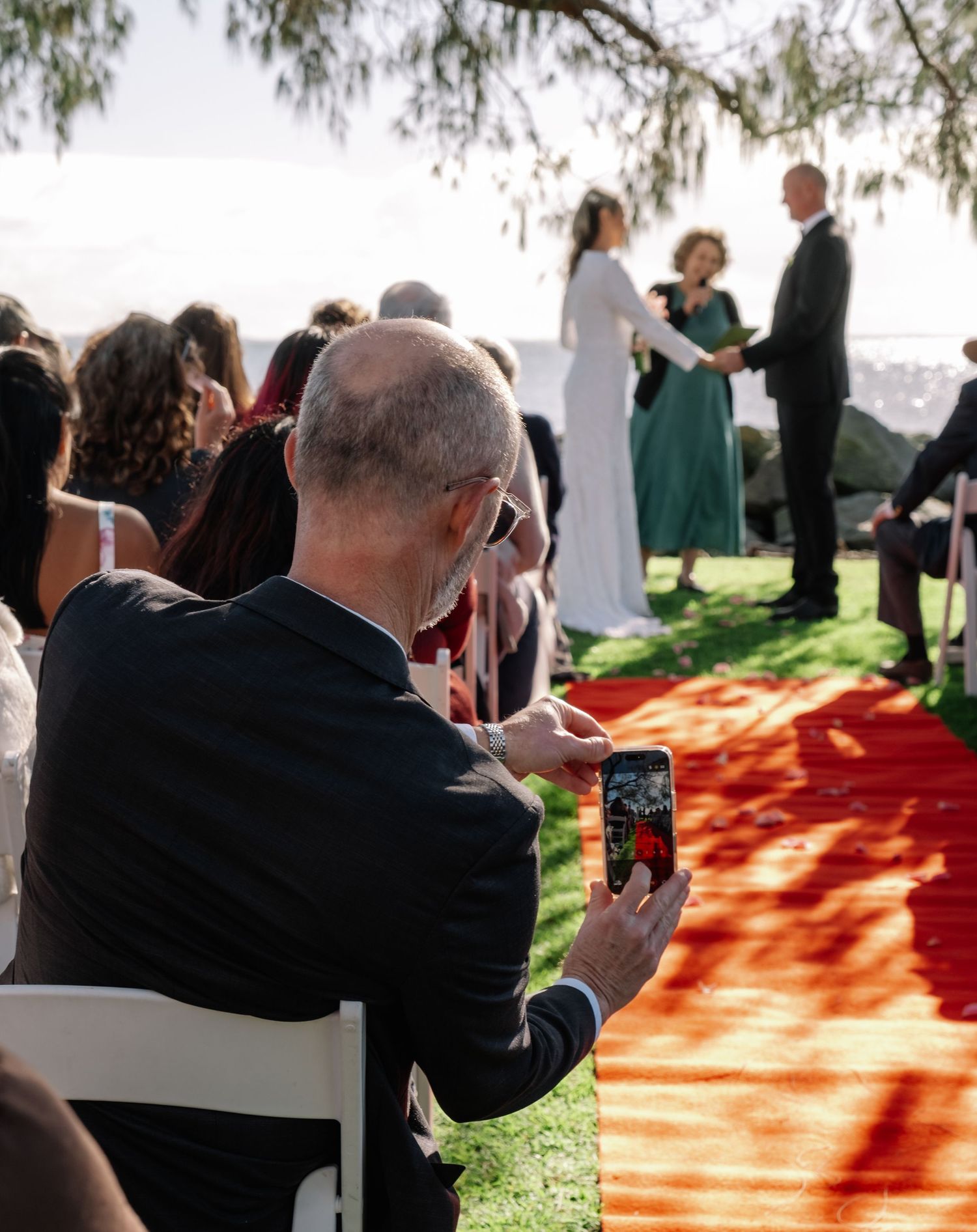 Wedding guest photographs ceremony on smartphone as couple exchanges vows by the ocean.