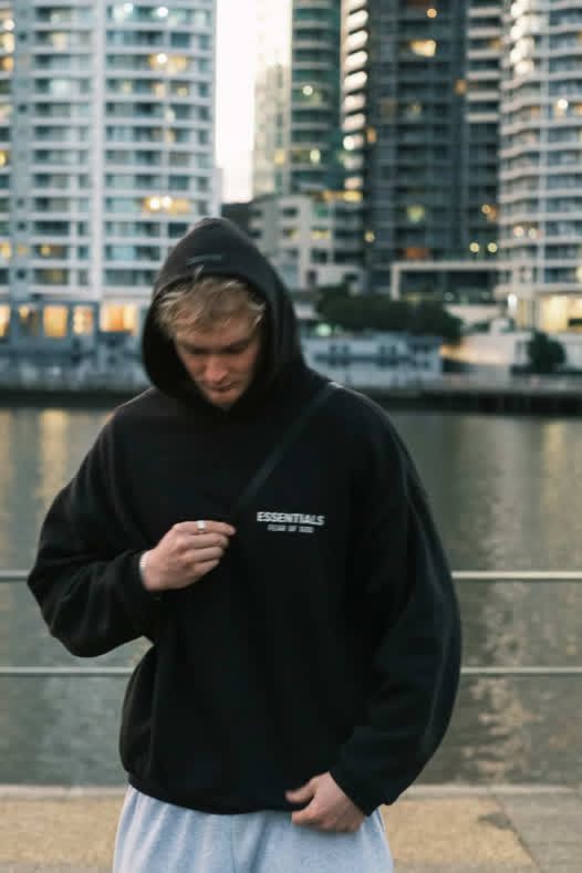 Man wearing black Fear of God Essentials hoodie standing by urban waterfront.