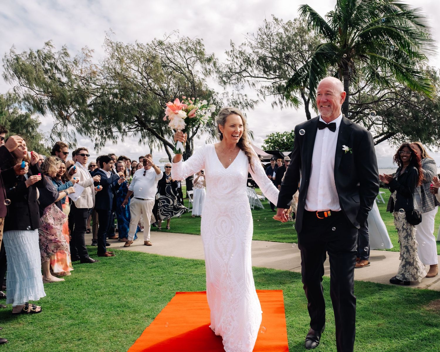 Smiling bride and groom walking down a red aisle runner surrounded by cheering wedding guests.