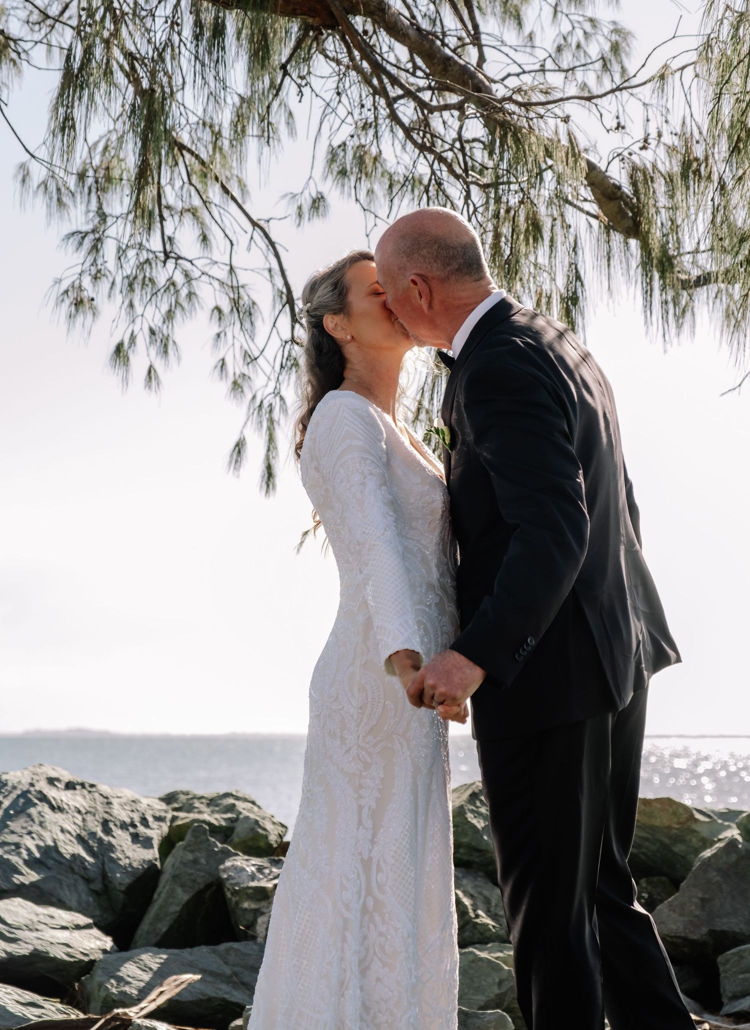 Bride and groom sharing a romantic kiss near rocky shoreline under coastal pine trees.